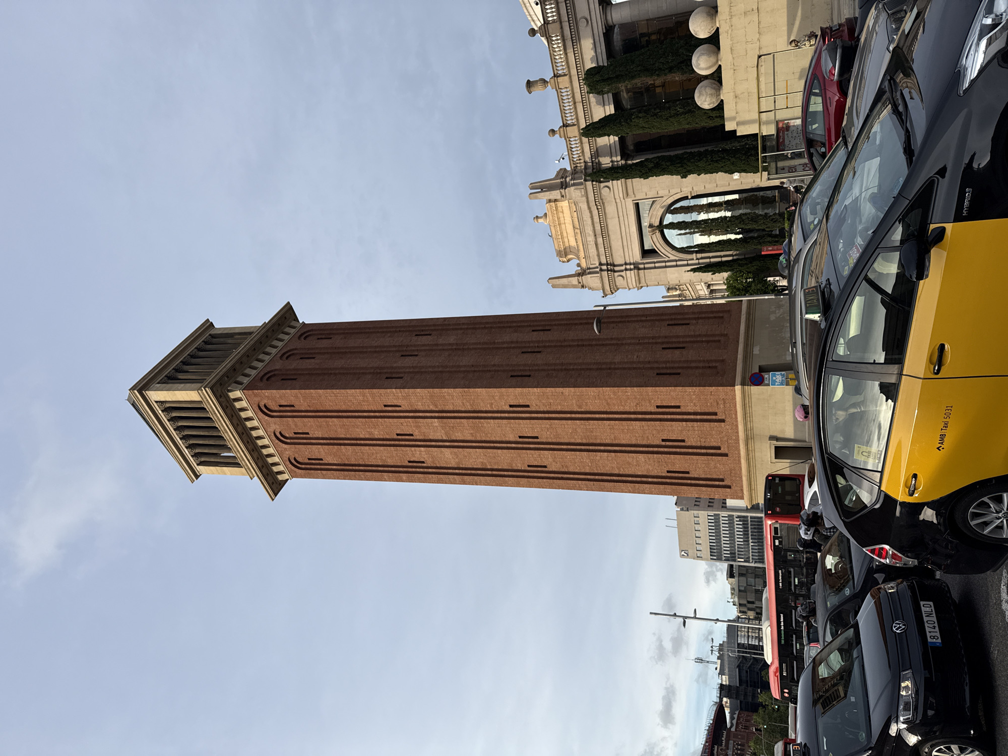 The image shows a tall brick tower, the Estació de França water tower in Barcelona, Spain. It is a tall, square structure with a decorative top. The foreground is filled with several parked cars, including a yellow and black taxi, as well as a red bus. The surrounding buildings and sky fill the background.