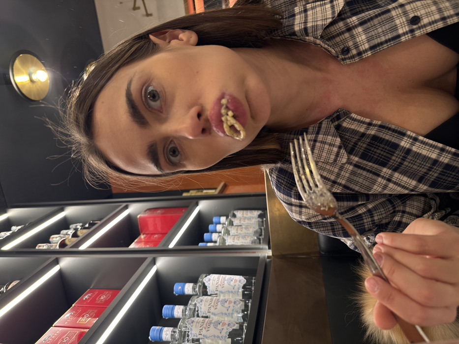 This image captures an indoor scene, likely within a modern restaurant or bar in Verona, Italy.
The primary subject is a female individual, appearing to be in her 20s or 30s, with medium-length dark brown hair featuring lighter highlights. She directly faces the camera with an exaggerated expression, her eyes wide open and her lips puckered, from which strands of light-colored pasta are visibly protruding. She wears a blue and white plaid button-up shirt, unbuttoned to reveal a black bra or bralette. Her right hand holds a clear plastic or glass fork, its tines showing residue of the same pasta and sauce.
The immediate background on the left features dark shelving units equipped with horizontal LED strip lighting. These shelves display multiple red boxes on higher levels and numerous bottles of "PANAREA VODKA" on lower shelves. To the right, a warm-toned wall (orange/brown) is visible, leading up to a dark ceiling from which a single brass-toned, downward-facing light fixture is suspended. A glimpse of dark upholstery, possibly part of a booth or banquette, is seen behind the woman on the left. A light-colored, possibly fur-lined object, is partially visible in the bottom-left foreground.