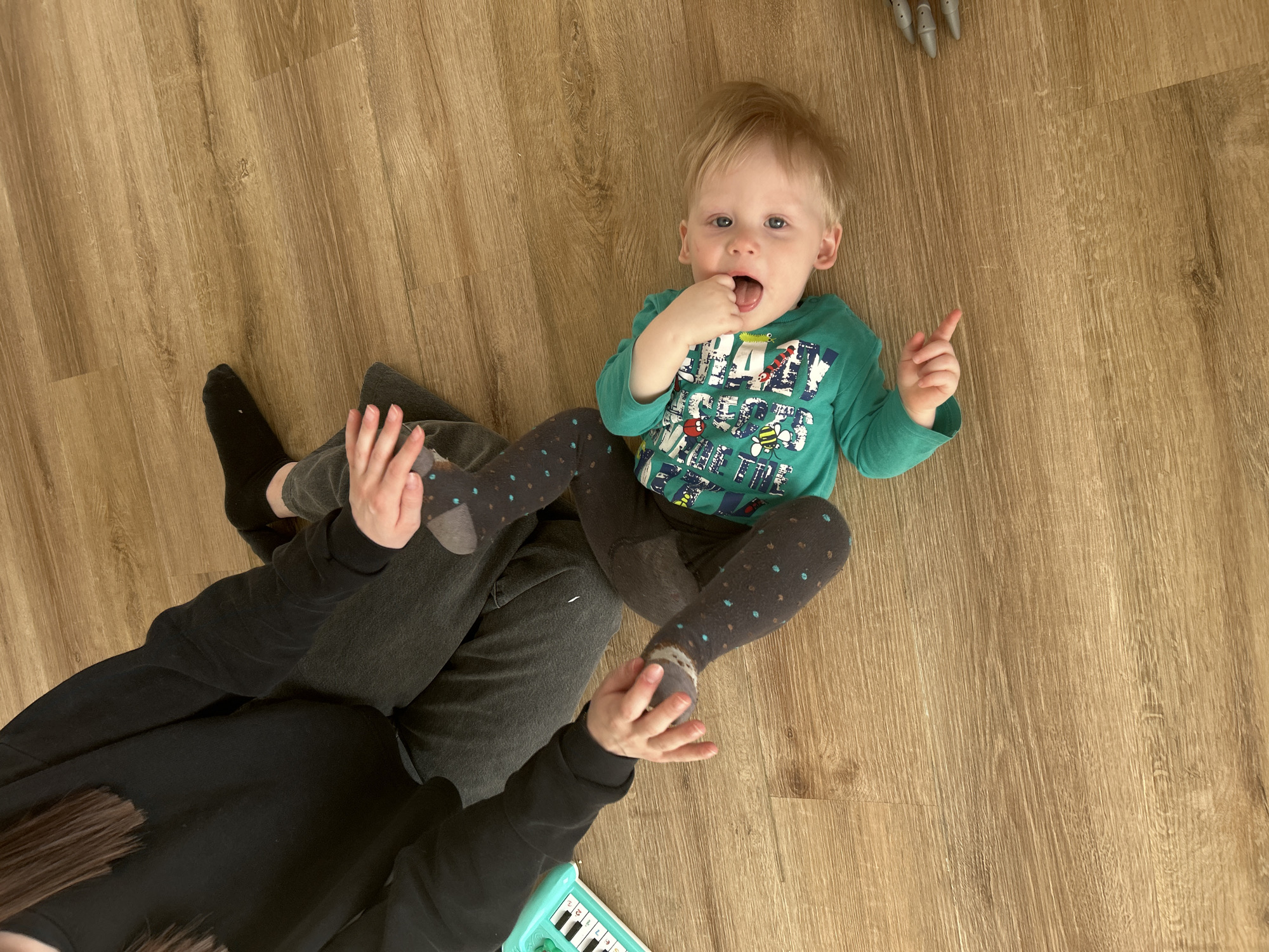 This image, taken in Chaikovsky, Russia, presents a top-down view of a toddler and an adult interacting on a light brown wooden floor.
The central subject is a fair-haired toddler with blue eyes, lying on their back and looking directly at the camera. They wear a green long-sleeved shirt featuring a graphic design with insects and visible text including "CRAZY". Their left hand has a finger in their mouth, while their right index finger points upwards. The toddler is also wearing dark tights with light-colored polka dots.
A partially visible adult is seated on the floor, wearing a dark long-sleeved top and dark pants or leggings with black socks. The toddler's legs are resting on the adult's lap, and one of the adult's hands is gently holding the toddler's foot.
To the bottom left of the frame, a small, light blue toy piano with white and black keys is visible. In the upper right corner, a metallic object with several finger-like protrusions can be seen on the wooden floor, possibly part of another toy.
The lighting suggests a bright, indoor daytime setting, likely a home. The scene depicts a calm, domestic moment, possibly of play or interaction between the adult and the toddler, who shows a curious or engaging expression.