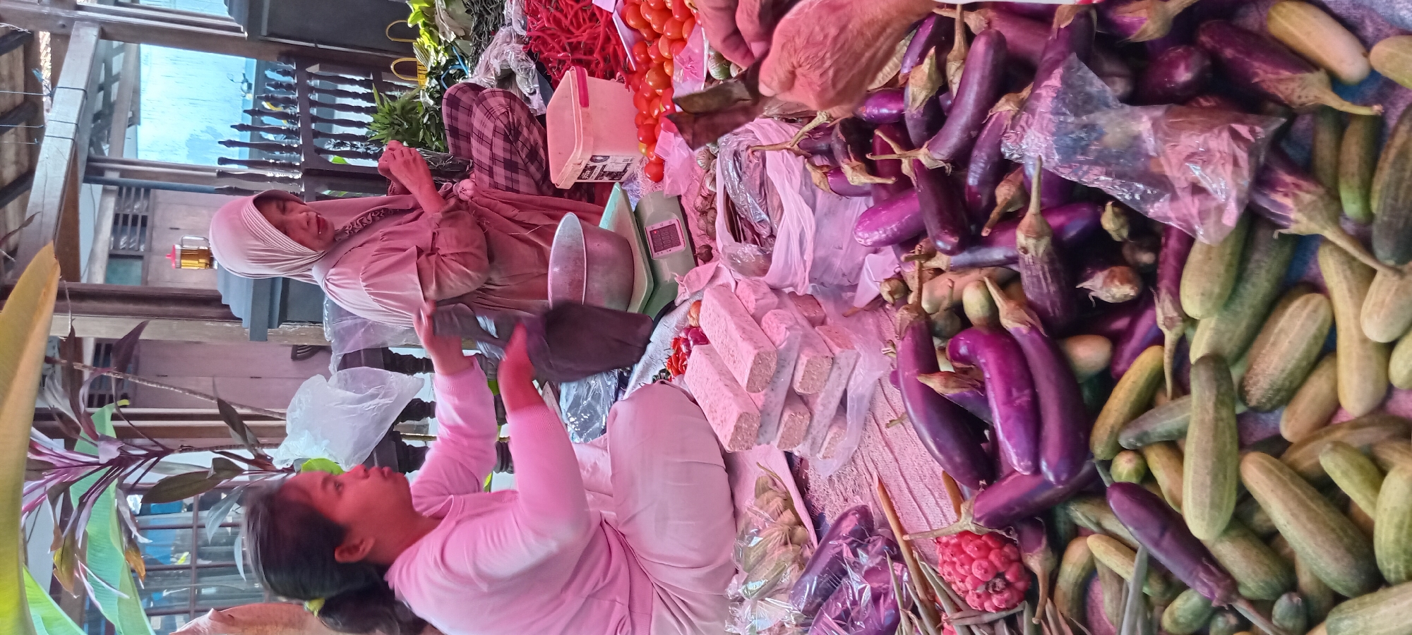 The image depicts a busy marketplace scene in Sindangwangi, Indonesia. Two women are engaged in transactions, surrounded by a vibrant display of fresh produce. On the left, a woman in a pink long-sleeved shirt and light-colored pants is seated, holding a black plastic bag and interacting with a scale. In front of her are several blocks of tempeh, along with other vegetables like chili peppers and shallots. To her right, another woman, wearing a light pink hijab and a patterned skirt, is seated and sorting through greenery.
The foreground is dominated by a large assortment of vegetables, including numerous dark purple eggplants, green cucumbers, and some red-skinned round vegetables that appear to be radishes or tomatoes. The produce is spread out on a woven mat, with some items placed in clear plastic bags. In the background, a rustic wooden structure and decorative railing suggest an outdoor or semi-outdoor market setting. A glass containing a beverage with a reddish tint is visible, alongside a plastic container and a metal bowl. The overall atmosphere is one of active commerce and the abundance of fresh food.