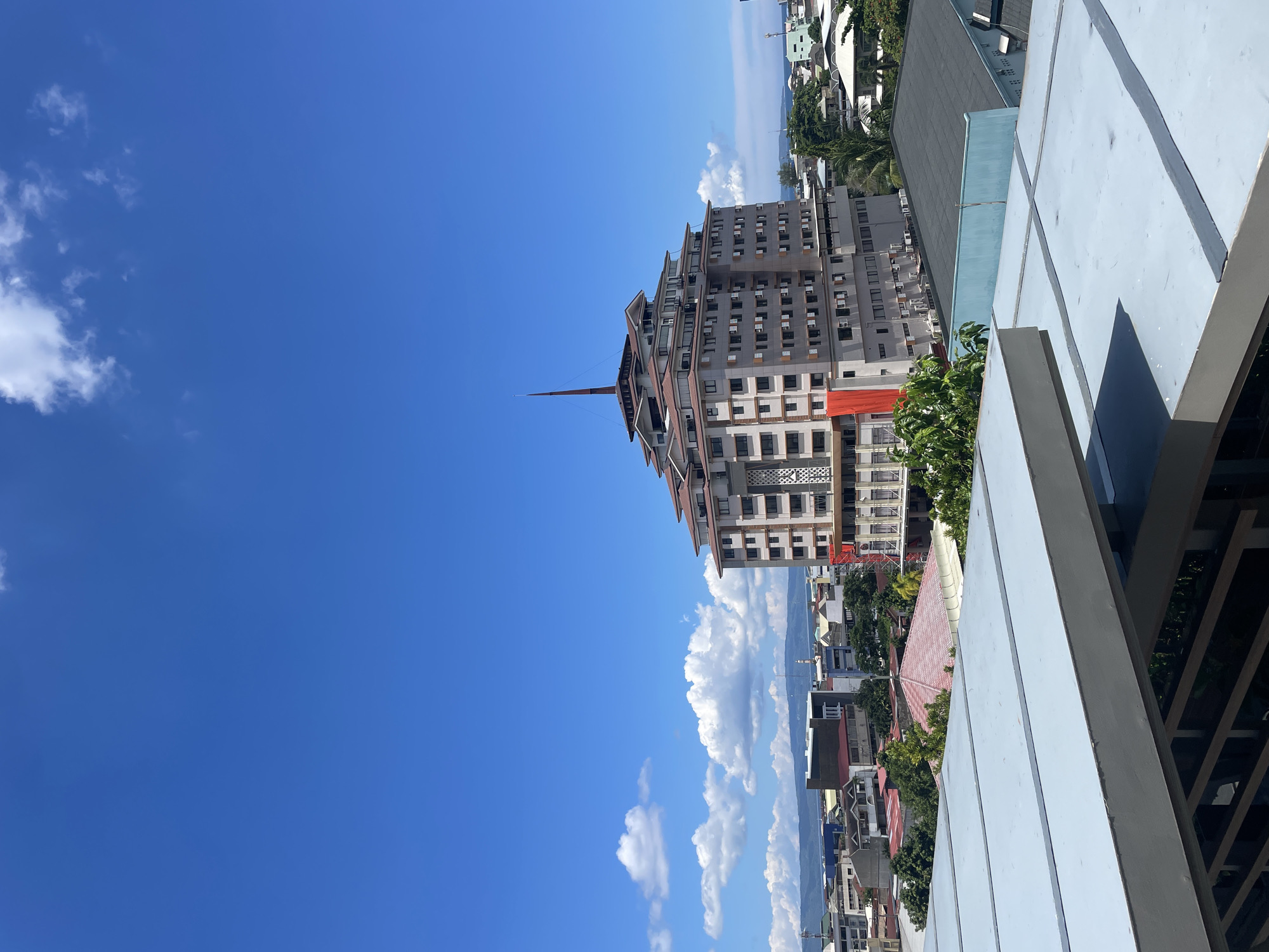 The image shows a high-angle view of a cityscape under a bright blue sky with scattered white clouds. In the center, a distinct, multi-layered building with a stepped design and a pointed spire dominates the skyline. The building's facade is a mix of gray panels and numerous windows, with a bright orange banner visible on one side. Surrounding this central structure is a denser area of buildings, featuring a variety of architectural styles and roof colors, including red-tiled roofs and flat, dark-colored roofs. Lush green trees are interspersed throughout the urban landscape. In the foreground, a portion of a modern, light gray structure with horizontal panels suggests a rooftop or elevated walkway, partially obscuring the view of the lower buildings. Beyond the city, a range of hills or mountains can be seen against the horizon, and a hint of the ocean is visible on the right side of the frame. The scene captures a typical day in a tropical urban environment, likely in Davao City, Philippines, characterized by dense development, vibrant green foliage, and clear, expansive skies.