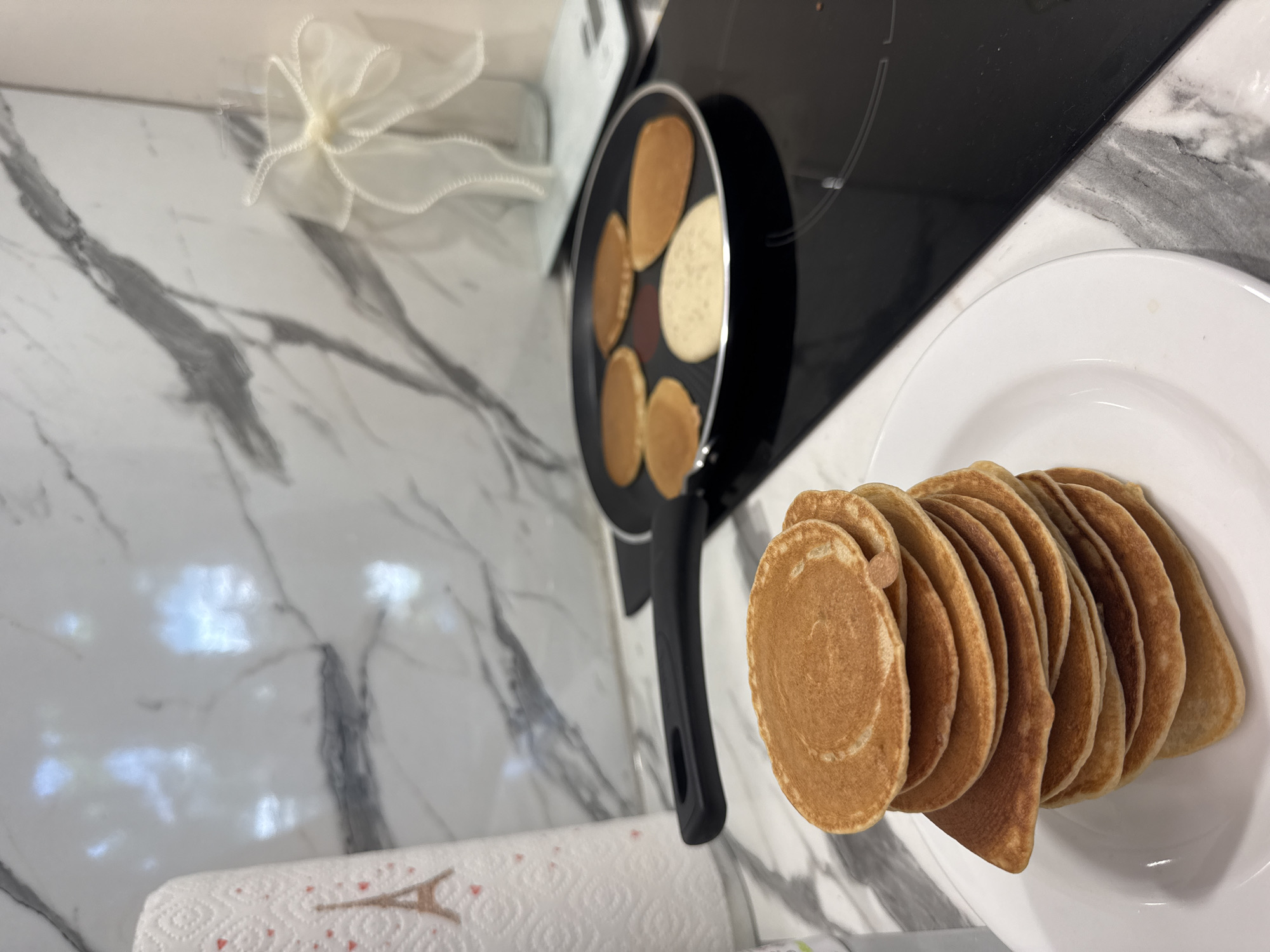 A stack of golden-brown pancakes sits on a white plate in a kitchen. In the background, a frying pan containing several pancakes is on a black induction cooktop. A roll of paper towels with an Eiffel Tower design is to the left of the pancakes. The countertop is marble-patterned. A decorative glass vase with a white bow is visible in the upper right corner. No people are present in the image, and there is no indication of the location being in Sofia, Bulgaria.