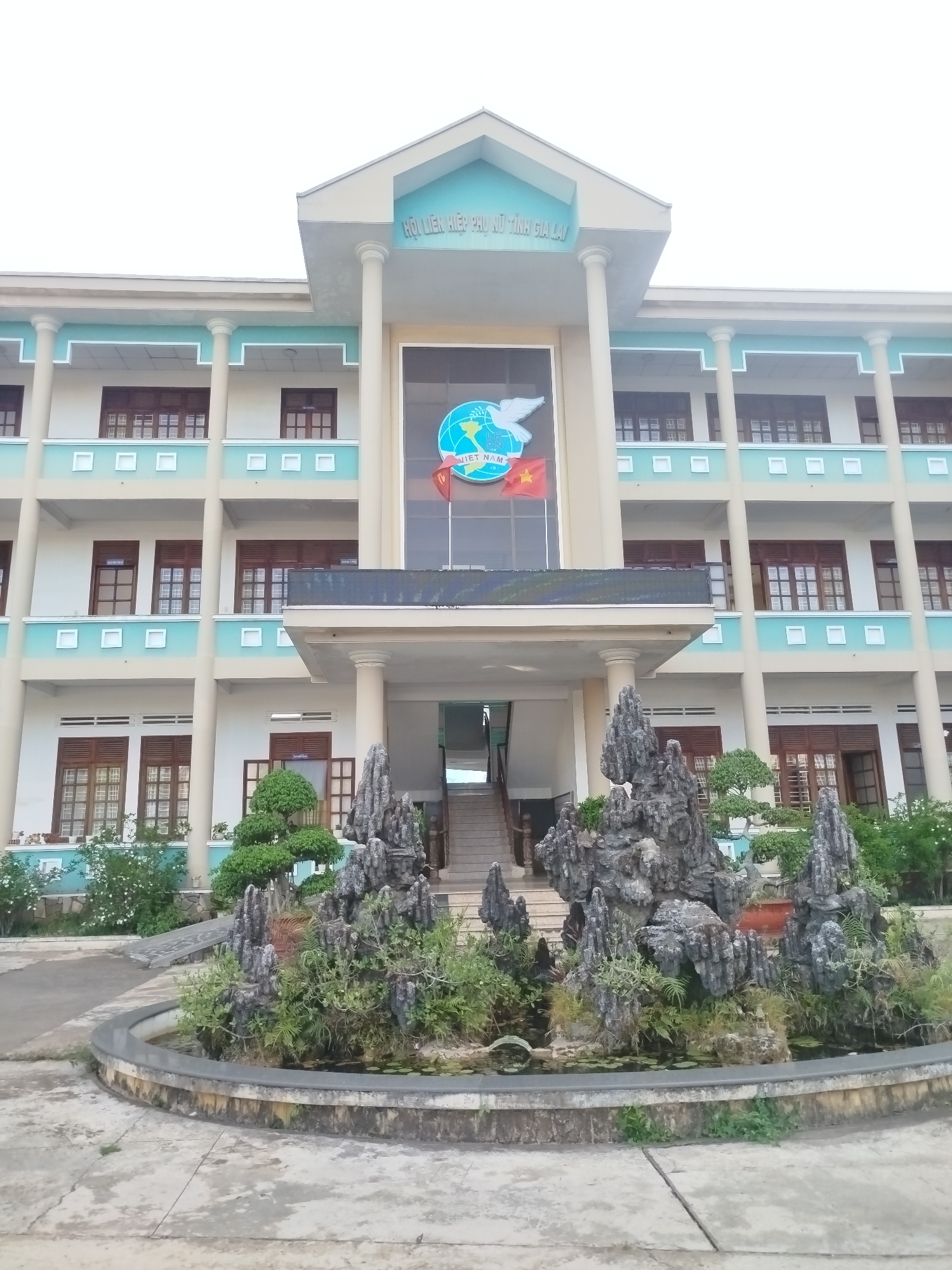 The image depicts the front exterior of a three-story building in Pleiku Ward, Vietnam. The building is characterized by a symmetrical design, with multiple rows of windows, cream-colored walls, and turquoise accents.
At the entrance, a water feature with rocks and plants occupies the foreground.
A sign with what appears to be the name of the organization is visible above the entrance, along with a logo and the Vietnamese flag. The building's architecture suggests it might be an administrative or educational facility.
The scene is set in daylight, with overcast skies casting a soft light on the surroundings.