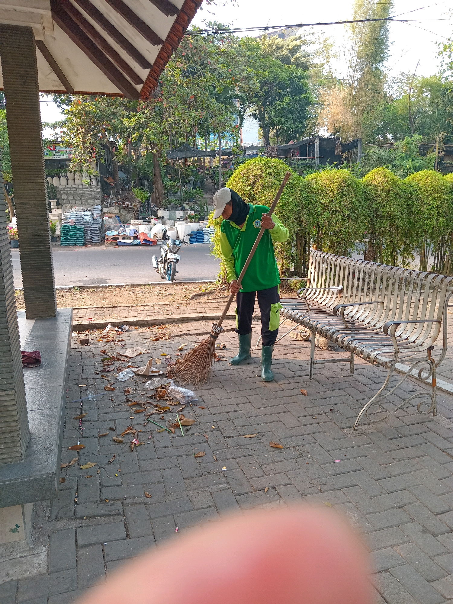 In Randuagung, Indonesia, a worker wearing a green long-sleeved shirt, black pants with green accents, and green boots is sweeping a cobblestone pathway with a broom. The worker also wears a black head covering and a white baseball cap. The pathway is littered with dried leaves and small pieces of trash. To the worker's right is a metal park bench. Behind the bench, a dense bamboo hedge creates a visual barrier. In the background, a road is visible with parked motorcycles and stacks of bagged materials. Trees and foliage are abundant in the surrounding environment. The scene appears to be outdoors during the day, with sunlight casting shadows.