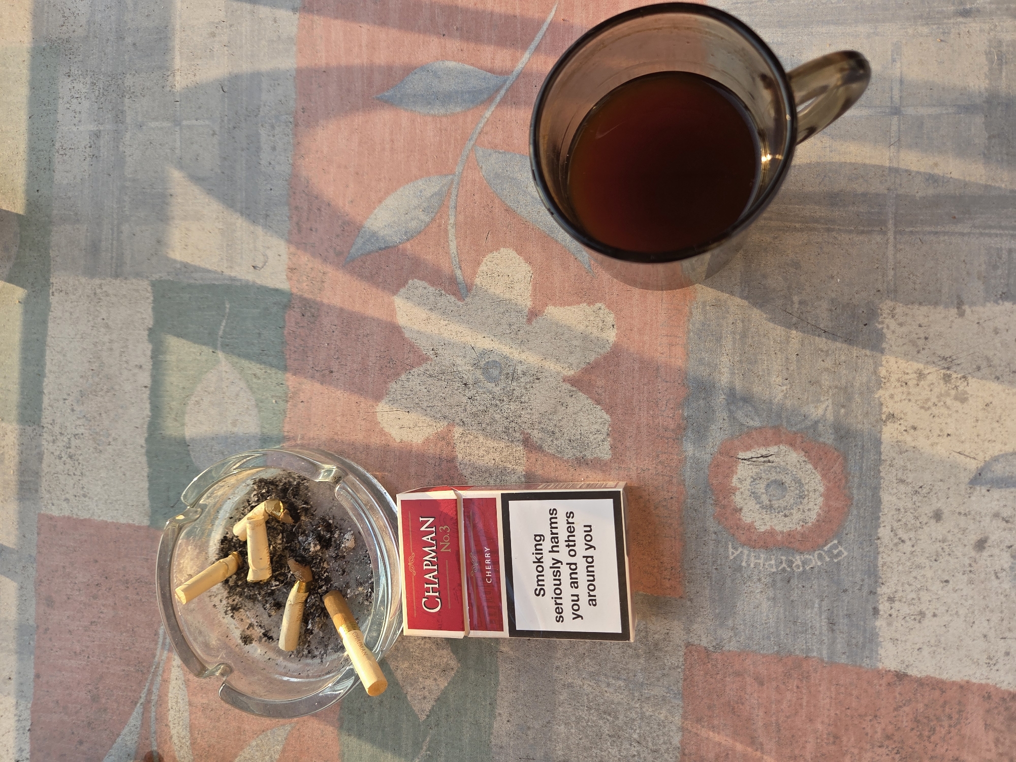 An overhead shot shows a glass ashtray filled with cigarette butts and ash, a pack of "CHAPMAN NO.3 CHERRY" cigarettes, and a mug of dark liquid placed on a tablecloth. The tablecloth has a pattern of faded floral designs and abstract shapes in shades of pink, red, gray, and white. Sunlight casts shadows of leaves and geometric shapes across the scene. The overall impression is of a person enjoying a moment of relaxation with cigarettes and a drink, possibly outdoors given the light and shadows.