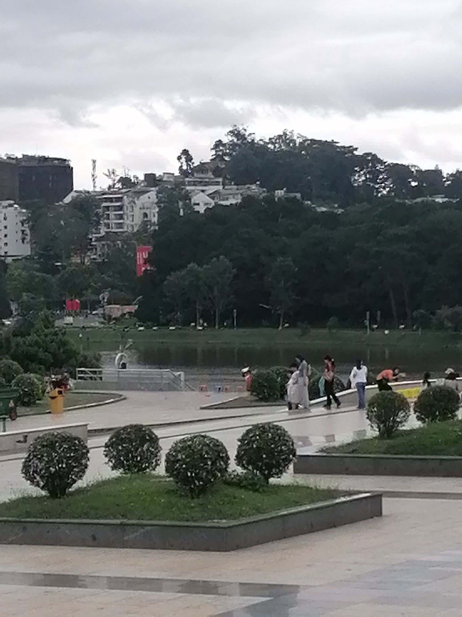 The image depicts a public park area in Da Lat, Vietnam, under an overcast sky. In the foreground, a paved pedestrian area with square planters containing shrubs is visible. Behind this area is a body of water, likely a lake or pond, with a grassy area beyond. Several people are walking along the path next to the water. Buildings and denser vegetation can be seen in the background, indicating an urban environment.