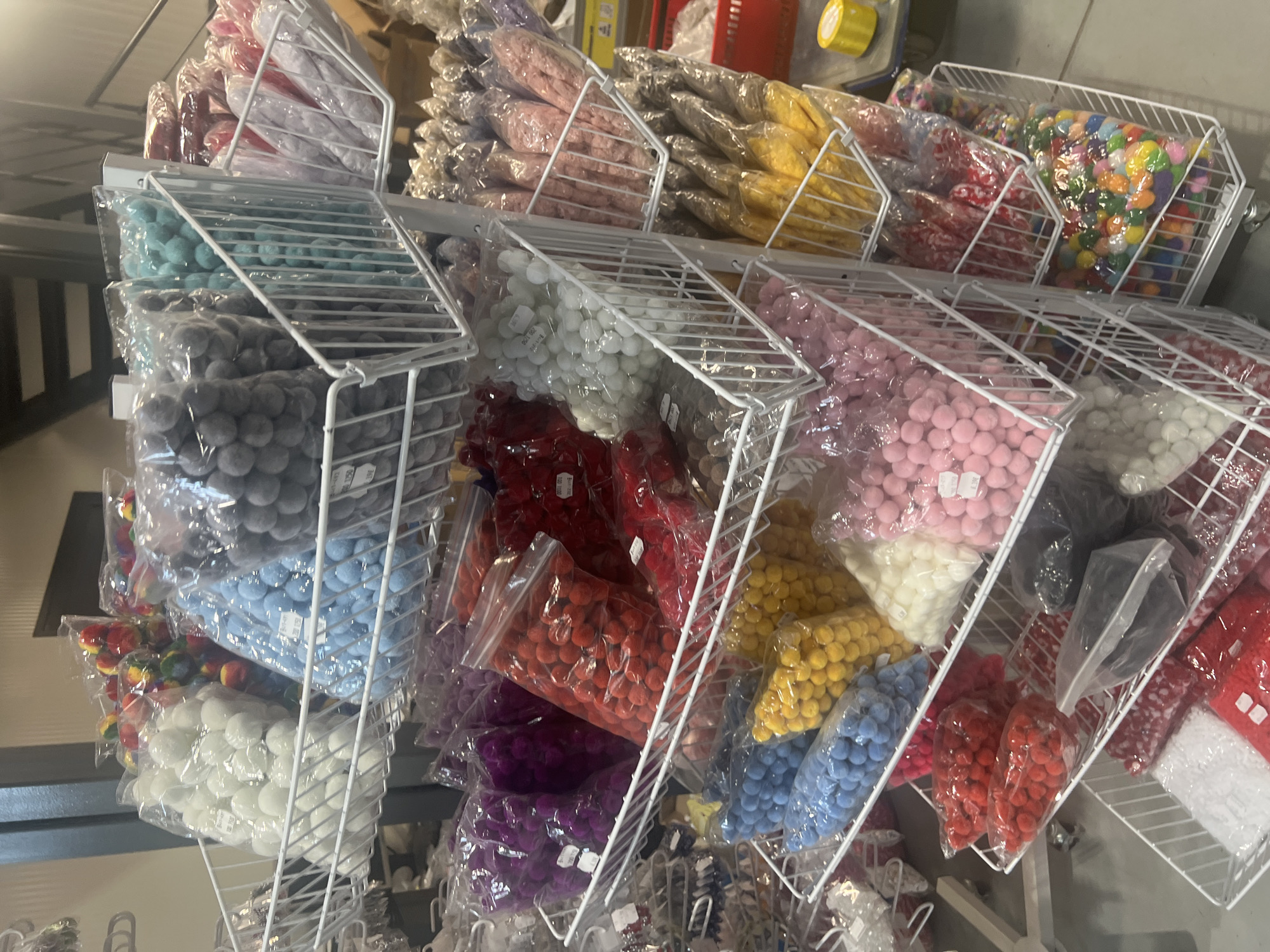 The image shows shelves of colorful pom-poms in plastic bags, organized by color in white wire baskets. The pom-poms appear to be for sale, potentially in a craft store or supply shop. The image was taken in Burgas, Bulgaria.