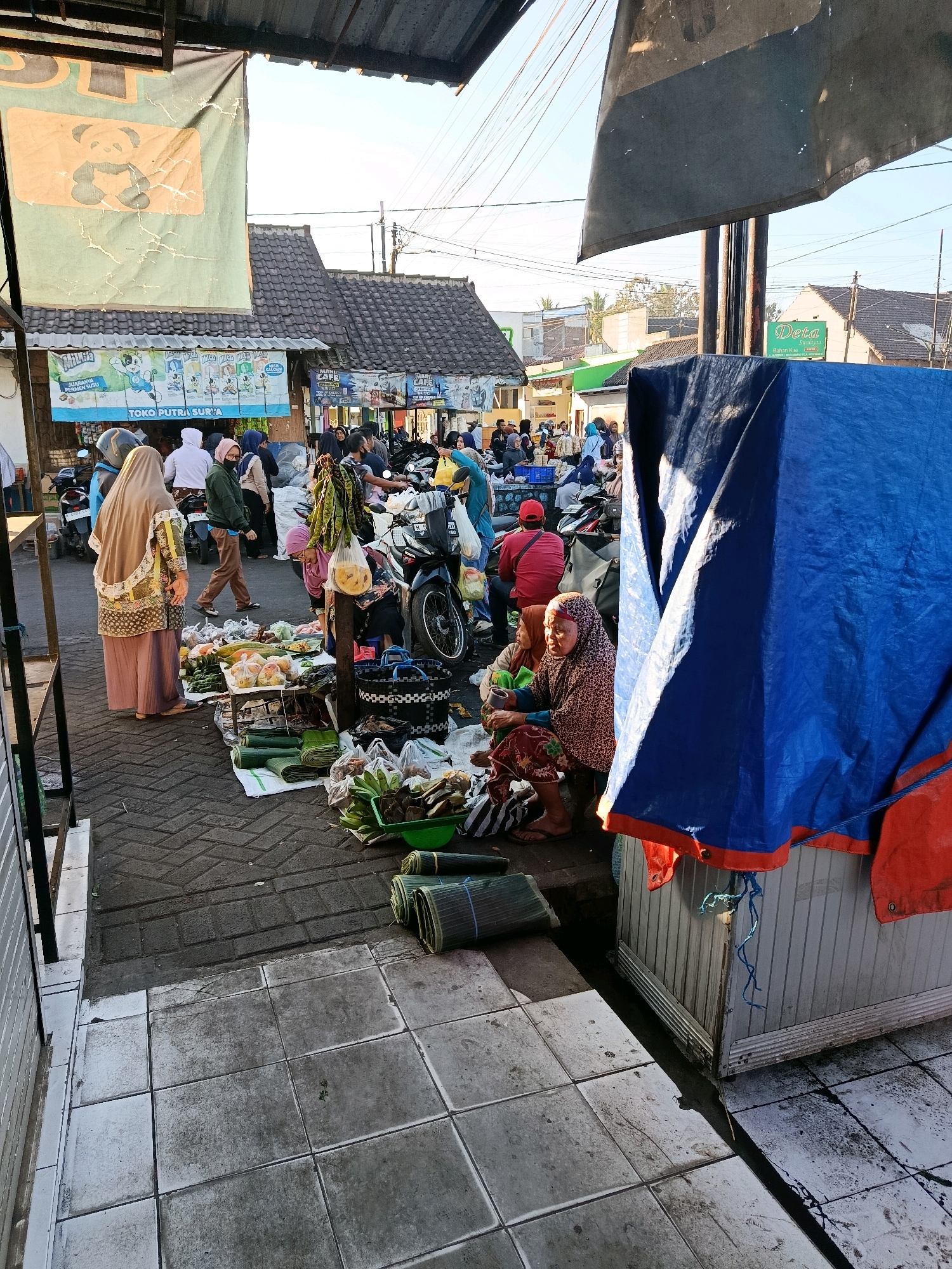 This image depicts a busy market scene in Bululawang, Indonesia. In the foreground, a variety of fresh produce, including bananas, cucumbers, and other vegetables, are displayed on the ground and on a low table. Several people are seen browsing and interacting with the goods. A woman wearing a light brown hijab and a patterned outfit stands to the left, facing away from the camera. To her right, a woman in a patterned headscarf and a dark patterned outfit is seated, looking towards the market. Rolled mats, likely for use as seating or packaging, are stacked on the ground.
In the middle ground, more market stalls are visible, with a variety of goods for sale, including what appears to be clothing and household items. Several motorbikes are parked, indicating common modes of transportation in the area. People are engaged in transactions and general market activity. Several vendors appear to be setting up or attending to their stalls. Above the stalls, various signs and banners are hung, some of which are faded. One prominent banner on the left features a cartoon panda.
The background shows the typical urban landscape of Bululawang, with buildings, rooftops, and utility poles visible under a bright sky. The overall impression is one of a vibrant local market during daylight hours, characterized by its bustling activity and the presence of fresh produce and various commodities. The image is captured from a slightly elevated perspective, looking down into the market.