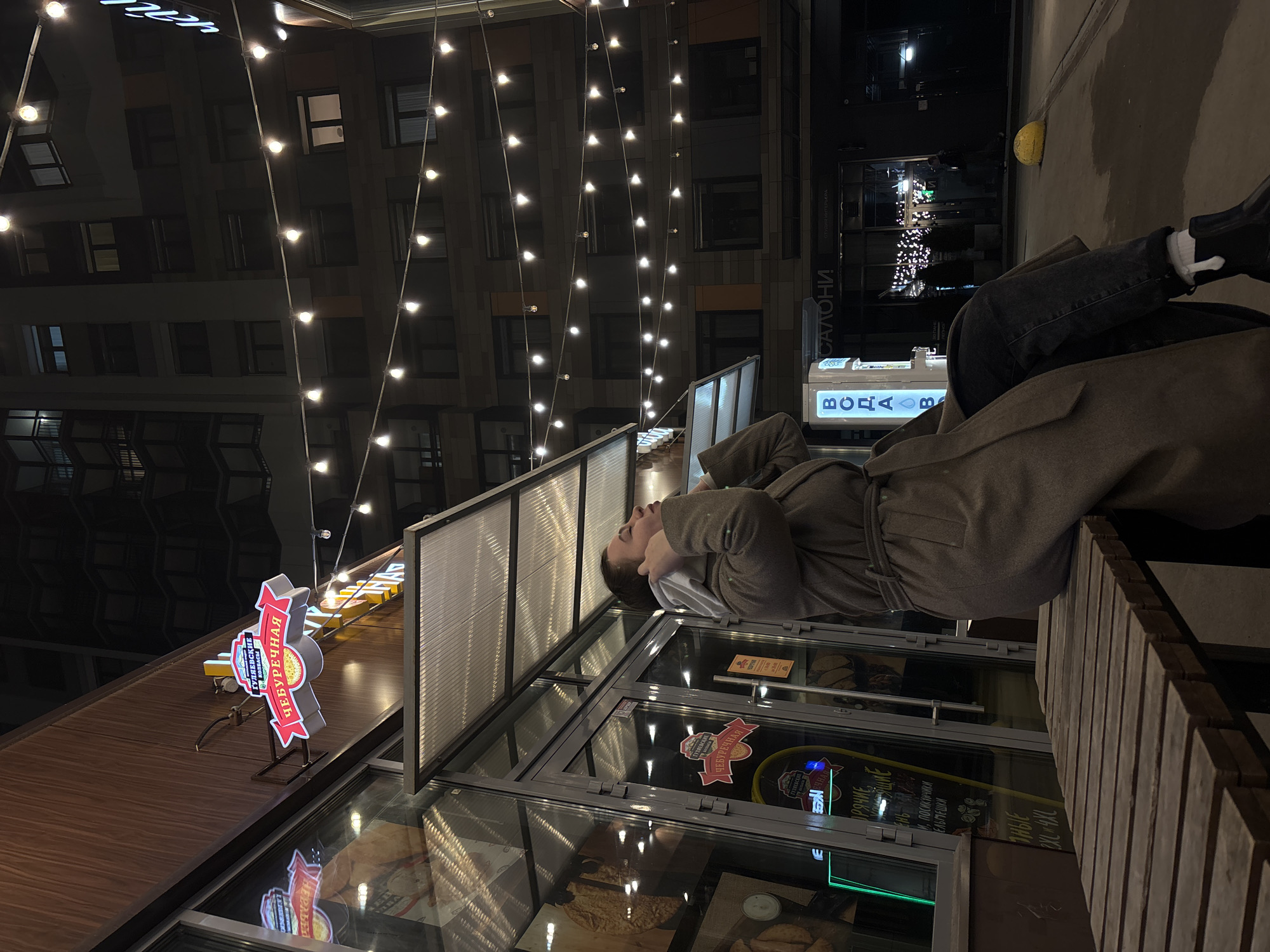 Here is a description of the image:
**People:** A person is sitting on a wooden bench, possibly resting their head near a food establishment. They are wearing a gray coat, dark jeans, and white socks with black shoes.
**Subjects and Objects:** The location appears to be an outdoor area next to a food establishment, indicated by the signage featuring words like "ЧЕБУРЕЧНАЯ" (Cheburechnaya), "ОРЯЧИЕ" (Hot), and "ГУЛЯЕВСКИЕ КОЛБАСЫ кол" (Gulyaevskie Kolbasy kol). The food establishment has a transparent display case with various food items inside. There are also decorative lights strung overhead.
**Actions and Interactions:** The person is sitting and appears relaxed, possibly observing the surrounding area or taking a break.
**Environment:** The photo seems to have been taken at night or during twilight, as evidenced by the darkness and the lighted buildings in the background. The environment includes buildings with multiple windows and lit signs.
**Additional Notes:** The food establishment is identifiable by the Cyrillic text "ГУЛЯЕВСКИЕ КОЛБАСЫ кол," indicating that the location is likely within Russia or a region where Russian is spoken. The overall atmosphere suggests a casual outdoor dining or rest area in an urban environment.