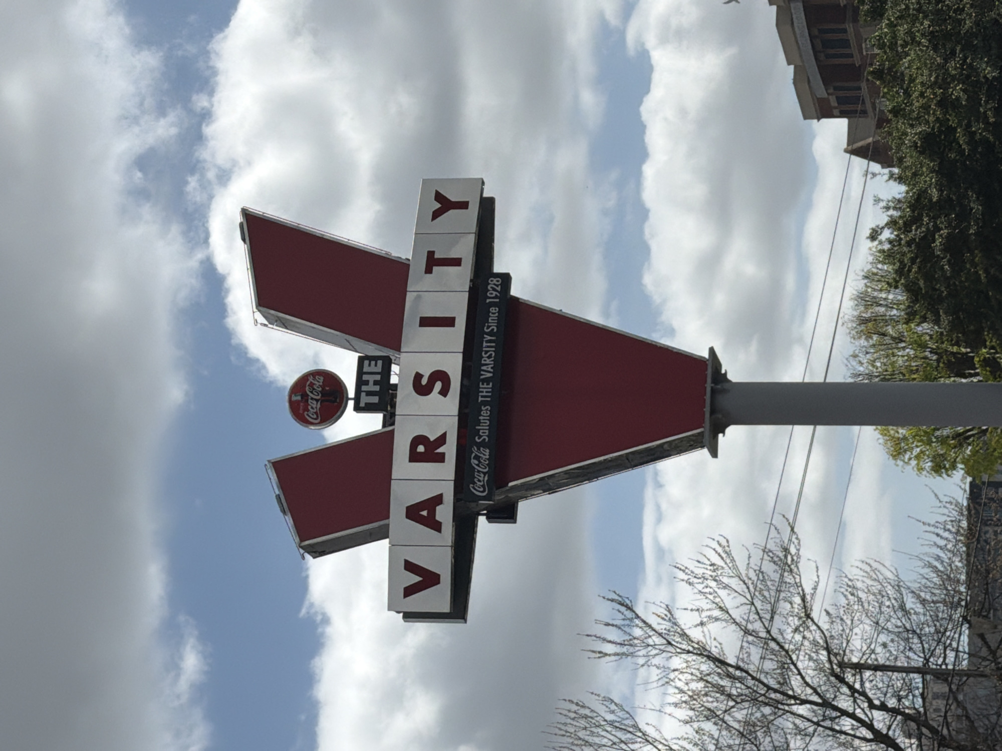 The image shows The Varsity, a famous fast food restaurant in Atlanta, Georgia, United States of America. The sign, a red and white structure with the word "VARSITY" in vertical block letters, stands prominently against a cloudy sky. A smaller Coca-Cola logo is affixed to the sign. The building and tree foliage in the background.