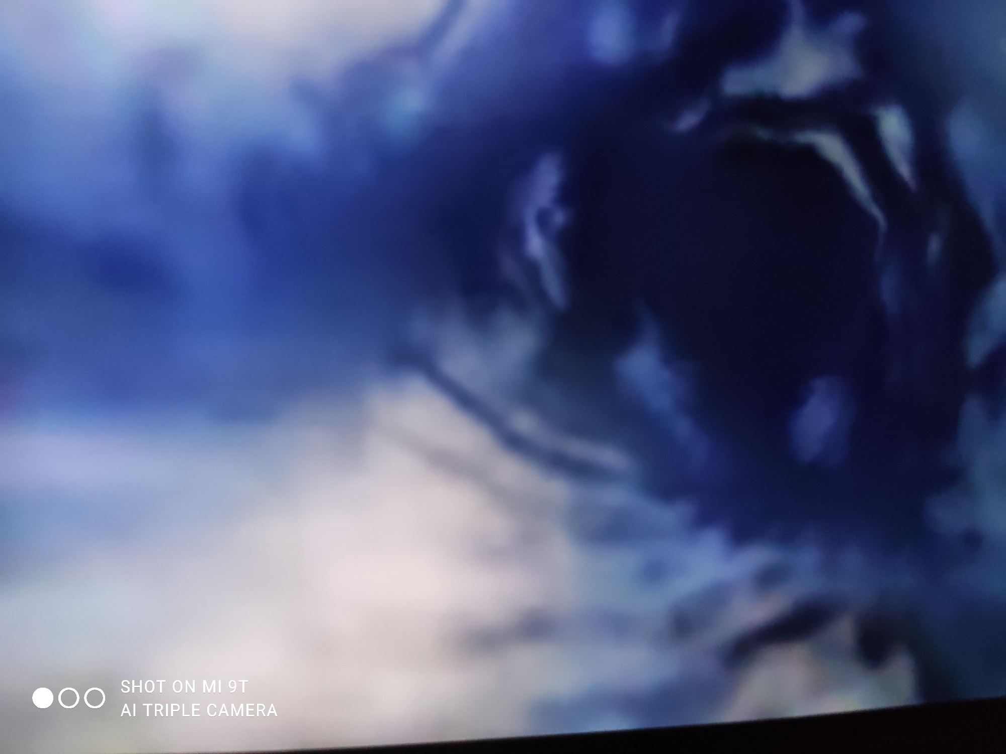 The image shows a blurry, abstract depiction of dark, swirling clouds or smoke, predominantly in shades of deep blue and white. The lighting is dramatic, with bright areas contrasting with deep shadows. The overall impression is one of turbulence and movement, possibly suggestive of a storm or an explosion. The bottom left corner of the image contains a watermark that reads "SHOT ON MI 9T AI TRIPLE CAMERA". The image is not of a specific location in the city of Khazar Raion, Azerbaijan, nor does it depict any discernible people, subjects, objects, actions, or interactions. It appears to be a digital image or screen capture with abstract visual content.