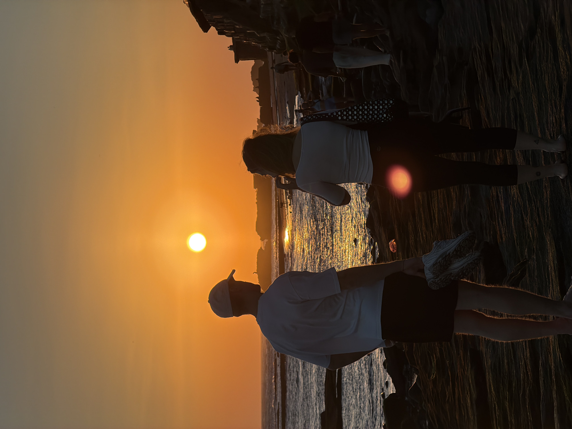 This image captures a vibrant sunset scene over a body of water, likely an ocean or large bay. The foreground features several people silhouetted against the bright, golden light of the setting sun, standing on what appears to be a rocky or paved shoreline.
Two main figures are prominent in the foreground. On the left, a man is seen in profile, wearing a light-colored t-shirt, dark shorts, and a baseball cap. He is looking towards the setting sun. To his right, a woman stands, also in profile, dressed in a light-colored long-sleeved top and dark leggings. A black bag with white polka dots is slung over her shoulder. A small tattoo is visible on her lower leg. Further to the right, another person's leg, clad in an athletic shoe, is partially visible. Additional indistinct figures are visible further back along the shore, also silhouetted.
The scene is set at dusk, with the sun, a distinct bright circle, positioned low in the sky, slightly to the left of the center. Its brilliant light creates a shimmering path across the water's surface. The sky is dominated by warm hues, transitioning from intense oranges and yellows near the sun to softer, hazy golds and light browns further away. The distant horizon shows faint silhouettes of land or structures.
The activity depicted is people leisurely observing and enjoying the sunset. The overall mood is peaceful and contemplative. No visible text is present in the image. The coastal setting and warm light are characteristic of scenic sunset views often found in locations such as Mengwi, Indonesia.