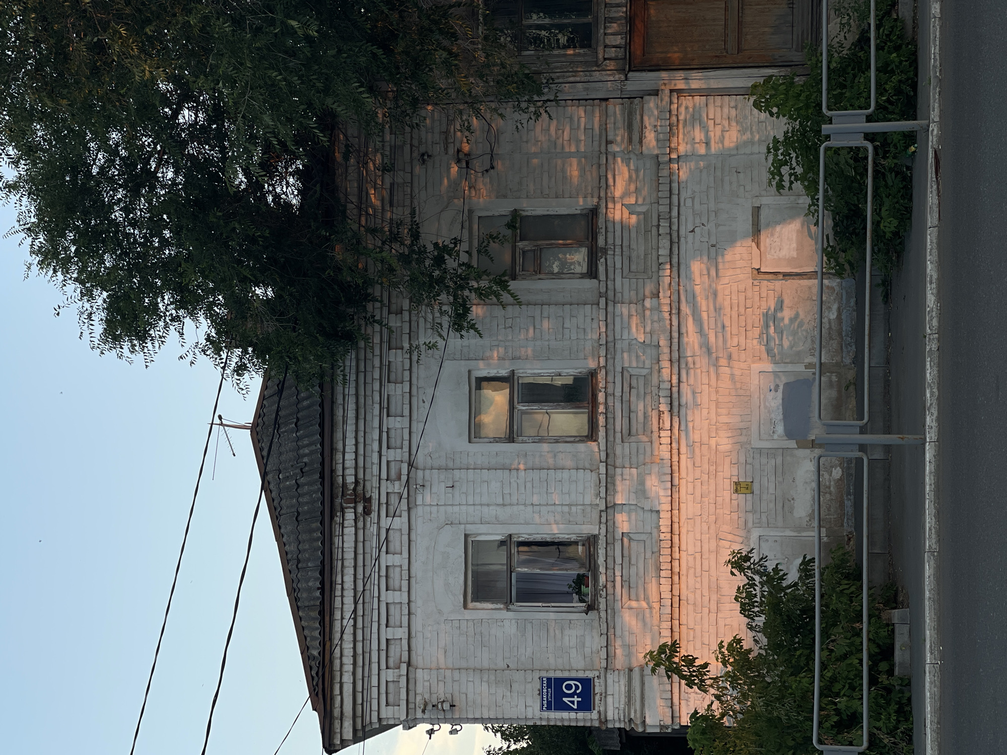The image depicts the facade of a two-story brick building, painted white, located at Rybakovskaya Street, house number 49, in Orenburg, Russia. The building exhibits signs of age and wear, with peeling paint and some discoloration on the brickwork. Sunlight, casting warm orange hues and dappled shadows, illuminates the left side of the building, highlighting the architectural details of the windows and decorative brickwork.
There are three windows visible on the upper floor, each with wooden frames and glass panes. One of the windows on the left has a small potted plant inside. A blue street sign with white lettering, displaying "РЫБАКОВСКАЯ 49," is affixed to the wall to the left of the first window. Below the windows, on the ground floor, are two recessed areas, suggesting former entrances or boarded-up openings. To the right of these, a weathered wooden door is partially visible.
A large tree with dense green foliage grows on the right side of the building, partially obscuring the upper stories and casting more shadows. In front of the building, a metal fence runs along the sidewalk, separating it from the street. Green bushes and smaller plants are growing behind the fence. The street in the foreground is paved, and a sidewalk runs alongside the fence. Overhead, power lines are strung across the frame against a clear, pale blue sky, with a television antenna visible on the roof. The overall impression is one of an old urban structure bathed in the soft light of late afternoon or early evening. No people are visible in the image.