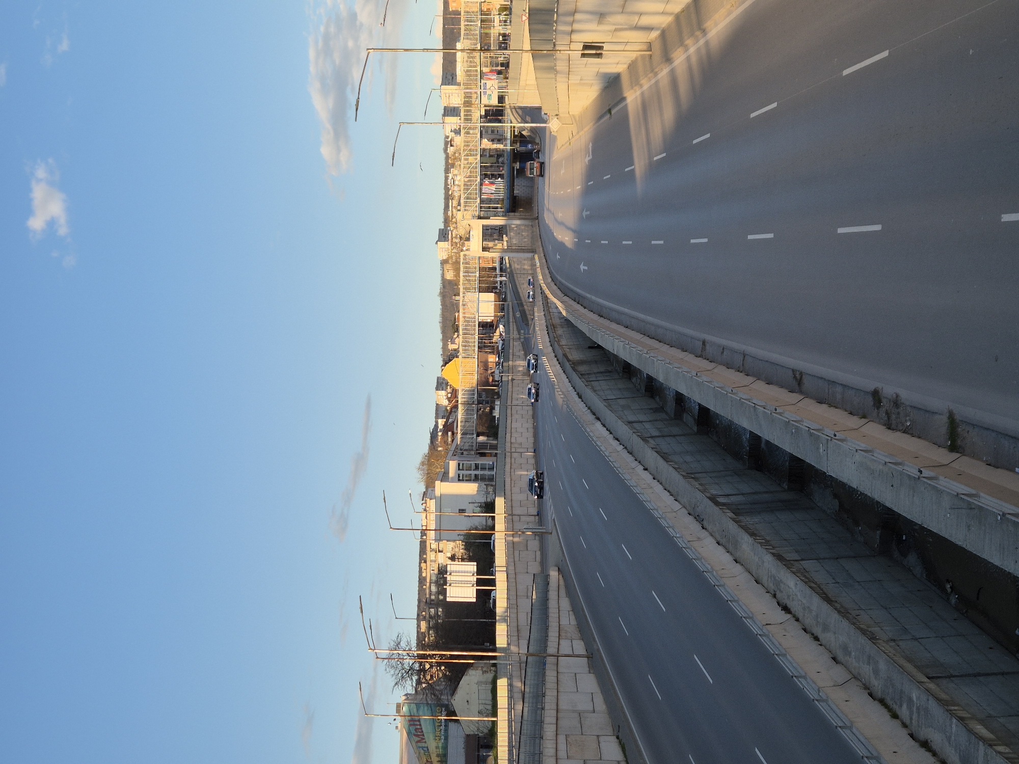 The image depicts a multi-lane highway running through a city, identified as Varna, Bulgaria. The highway is elevated with concrete barriers on either side. There are a few vehicles visible. Adjacent to the highway is a cityscape including buildings of varying heights and colors. Streetlights line the highway. A blue sky with some clouds is visible above the city. The image appears to be taken from an elevated position.