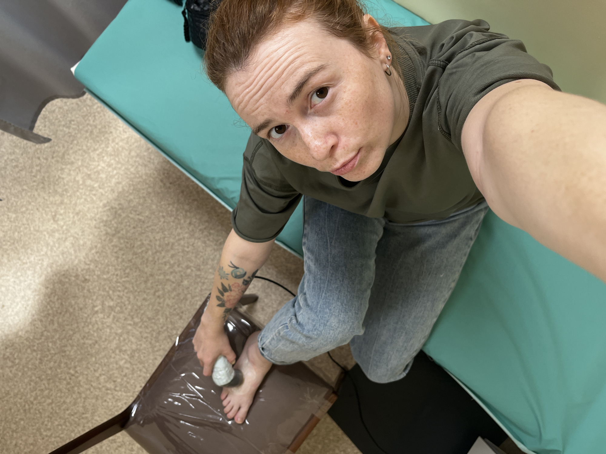 A high-angle selfie captures a woman with reddish-brown hair, pulled back, and a face adorned with freckles, looking directly at the camera with a neutral expression. She wears a dark green long-sleeved shirt and light blue jeans. Visible on her left forearm are several floral or nature-themed tattoos.
The woman is engaged in a foot care activity, holding a light-colored, textured object—possibly a pumice stone or an electric foot file—in her left hand, which she is applying to her right foot. Her right foot rests on a low, dark brown stool or table that is covered with a clear protective plastic sheet.
The setting appears to be an indoor room, possibly a treatment area or a clinic, suggested by the teal-colored examination table or bench positioned behind her. The floor is covered with a light brown, speckled carpet. A black wire is visible on the carpet near her left leg. The overall scene suggests a personal care or wellness routine, potentially performed in a professional or semi-professional environment. There is no visible text in the image.