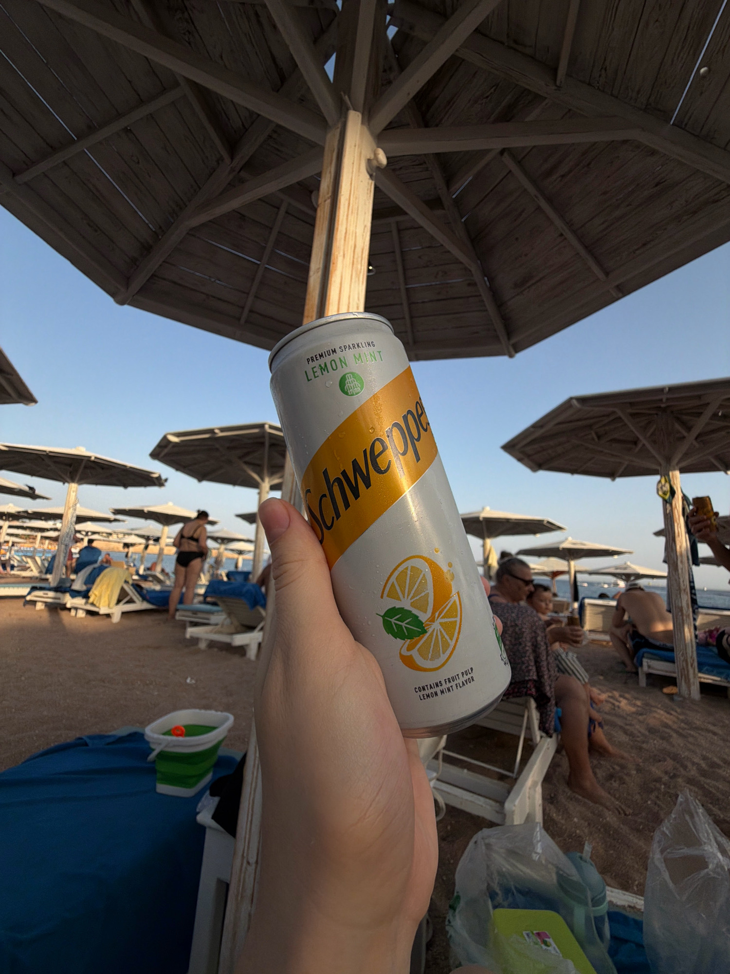 A person's hand holds a can of Schweppes Lemon Mint sparkling drink on a beach in Sharm El Sheikh, Egypt. The background shows beach umbrellas, lounge chairs with people relaxing, and the sea under a clear sky. The can has condensation on it. In the foreground, there is a blue beach mat and a collapsible bucket with a red object inside. The overall scene depicts a relaxed day at the beach.