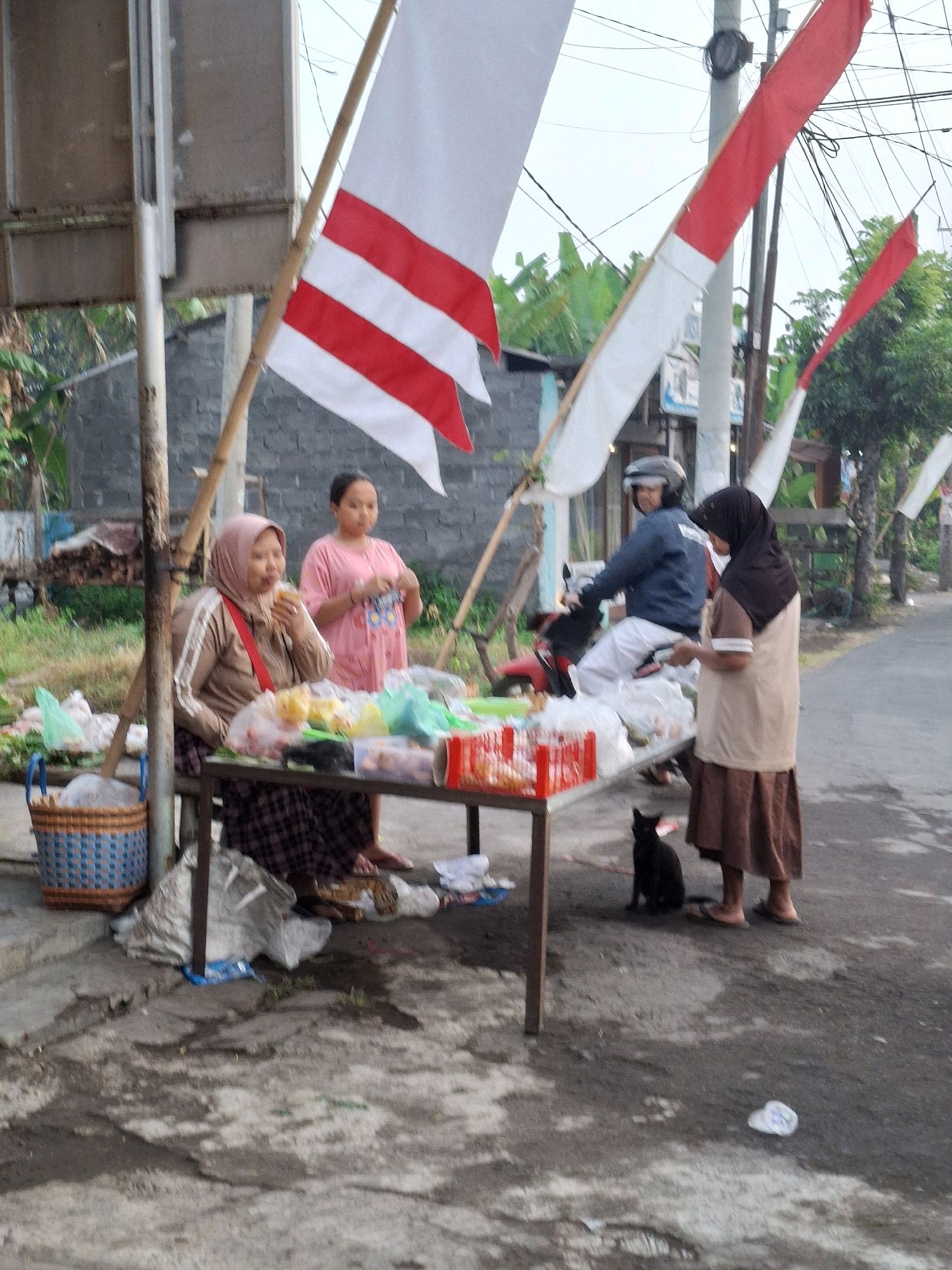 The image shows an outdoor scene in Lumajang, Indonesia, with several people and objects gathered around a table. Dominating the scene are two large Indonesian flags, red and white, draped on poles. On the left, a woman wearing a headscarf and a light brown jacket is seated at a table, eating. To her right, a younger woman in a pink shirt stands, holding something in her hands. A man wearing a helmet and a dark blue jacket is seated on a motorcycle behind the table. To the right of the table, a woman in a dark headscarf and a light brown and beige outfit stands, facing away from the camera. A black cat is seated on the ground near the table. The table is laden with various items, including plastic bags filled with goods, a red plastic crate, and some food items. A woven basket is placed on the ground to the left of the table. The ground appears to be paved with concrete and is wet in places. In the background, there is a brick wall, some trees, and electrical wires overhead, suggesting a semi-urban or roadside setting. The overall atmosphere suggests a market or roadside stall, possibly during a national celebration given the prominent flags.