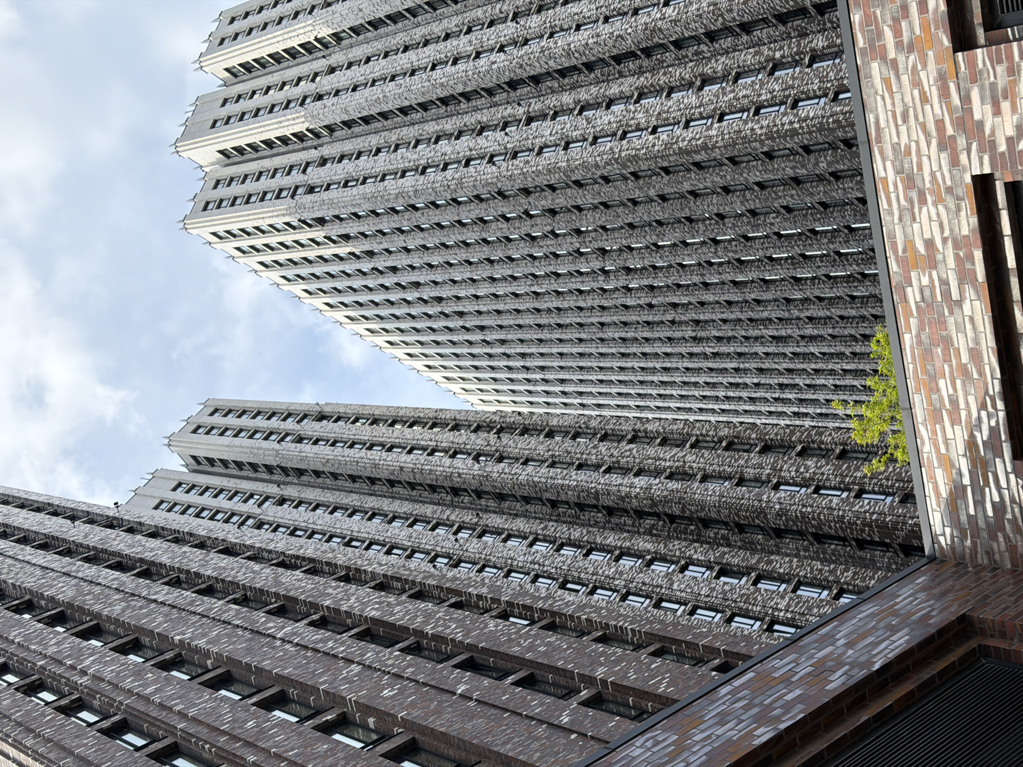 The image shows an upward-angled view of two tall buildings, likely in a dense urban environment. The buildings are characterized by a repetitive pattern of windows and horizontal lines, creating a sense of height and scale. The building in the foreground has a darker, brick-like façade, while the one behind it is lighter in color. A third building, partially visible on the right, has a multicolored brick pattern. A small amount of green foliage is visible. The sky is cloudy. No people are visible in the image. It is unlikely that this image is in Khimki, Russia, based on the architectural style depicted.