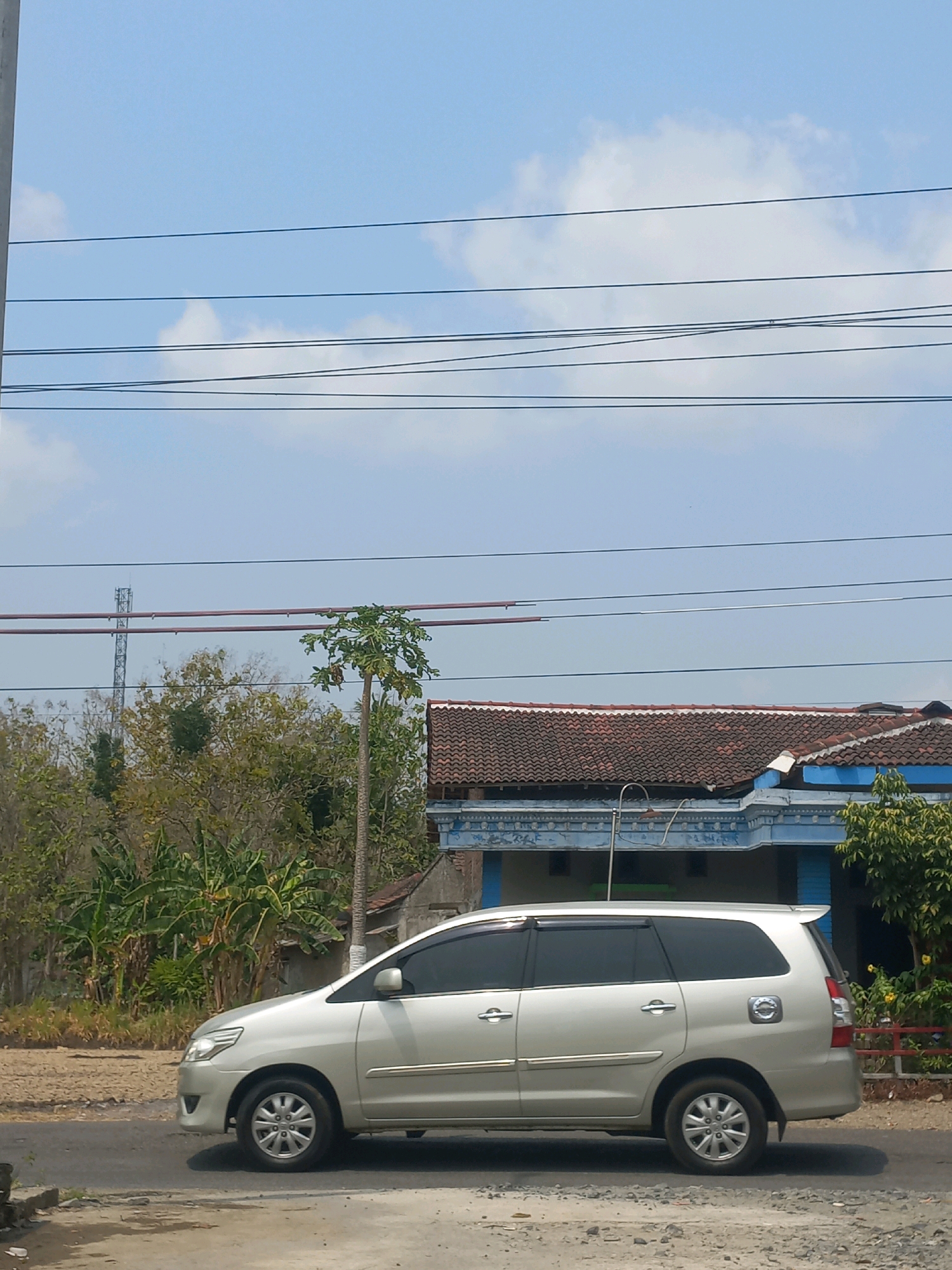 A silver minivan is parked on the side of a road in Baturetno, Indonesia. The vehicle is facing towards the left of the frame, and its doors are closed. Behind the minivan, there is a blue and white building with a terracotta tiled roof. Several trees and vegetation are visible in the background, including banana trees and a tall, slender tree with large leaves. A cell tower can be seen in the distance, to the left of the building. Power lines are strung across the upper portion of the image against a bright blue sky with scattered white clouds. The road surface appears to be asphalt, with some gravel and dirt on the side. The overall scene depicts a typical roadside view in a rural or semi-rural area of Indonesia.
