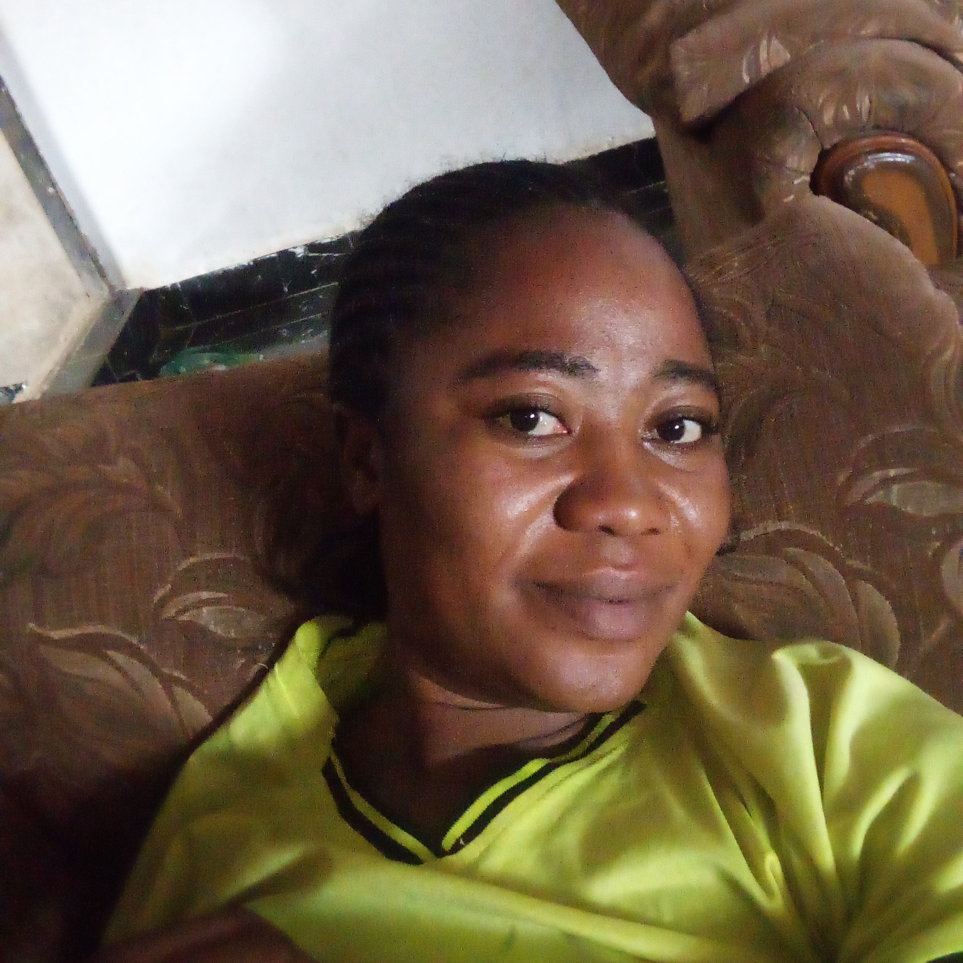 A dark-skinned woman with braided hair is centered in the frame. She wears a light green shirt with black stripes at the collar. She is reclining on a brown patterned couch. The background includes a wall with a white upper portion and a dark lower portion. The image appears to be taken indoors. The location, Goni Gara, Nigeria, is a detail unrelated to the visual content of the photograph.
