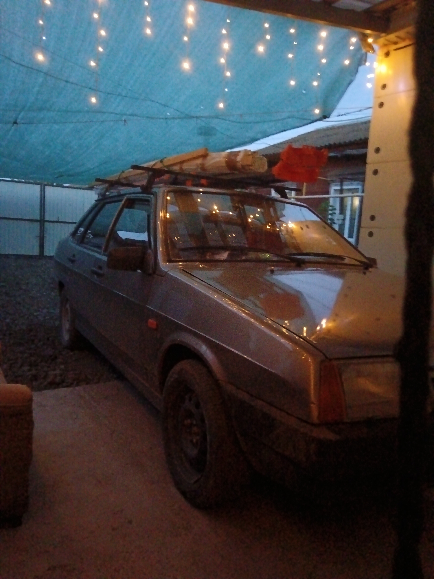 A vintage, grey four-door sedan, possibly a Lada, is parked under a blue tarp draped with strings of yellow lights. The car is loaded with wooden planks on its roof rack. The scene appears to be an outdoor space, possibly a yard or a private parking area, with gravel on the ground and a fence visible in the background. A portion of a building with white siding and windows is also present. The lighting suggests it is evening or dusk. The location is specified as Sorochinsk, Russia.