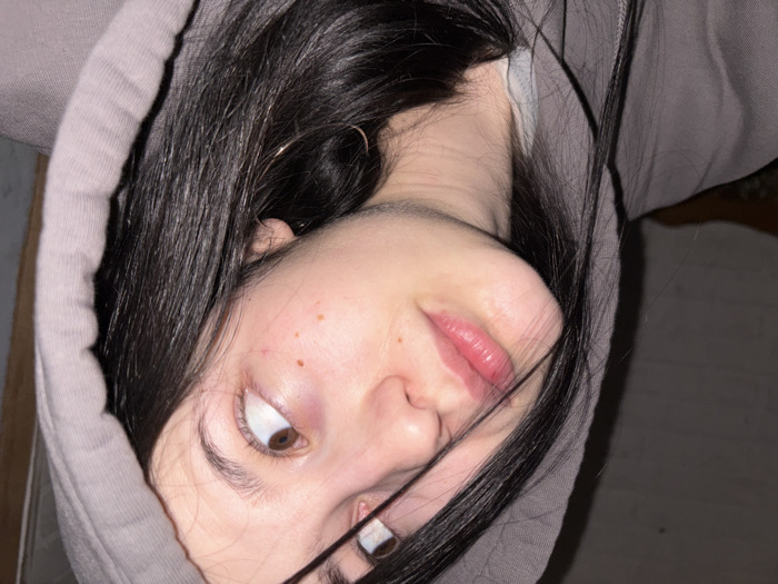 A young woman's face is angled upward, framed by dark hair and a grey hoodie. Her brown eyes gaze to the side, and a silver ring adorns a finger near her face. She has fair skin with freckles, and her lips are a natural pink. The background appears to be a dimly lit room, with a glimpse of a wooden door or wall visible.