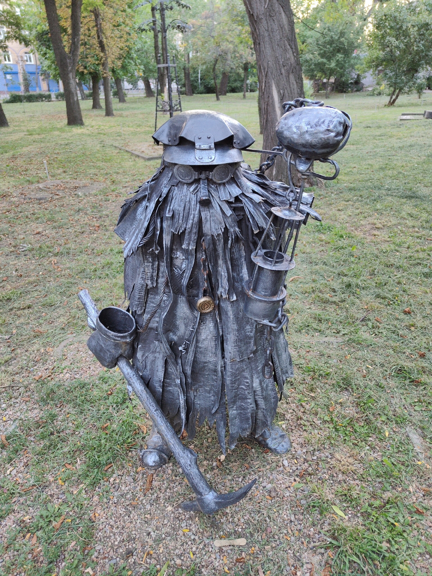 This image shows a metal sculpture of a miner in a park-like setting. The sculpture is made of dark, textured metal, giving it a rough and weathered appearance. The miner is depicted wearing a helmet with goggles, a tattered cloak-like garment, and boots. In its right hand, it holds a lantern, and in its left, it grips a pickaxe. The sculpture stands on a grassy area with scattered gravel and fallen leaves. In the background, trees with autumn foliage are visible, along with a building with blue-painted sections and windows. The overall impression is that of an outdoor art installation in a public space.