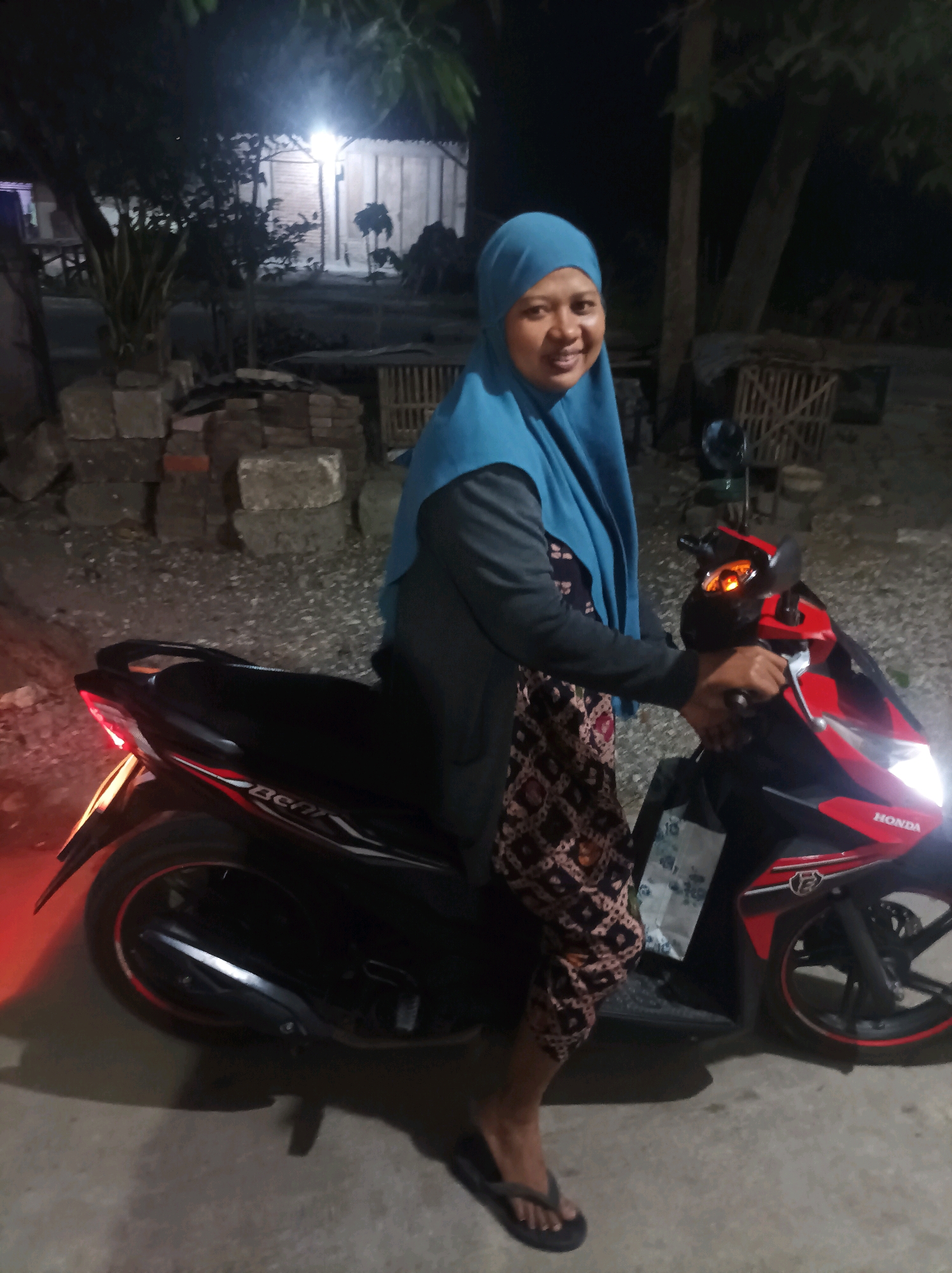 A woman in a teal hijab and patterned pants is seated on a black and red Honda scooter at night. She is smiling at the camera. The scooter's headlights and taillights are on. The background is dimly lit and appears to be an outdoor area with trees, a wooden structure, and a pile of bricks. The ground is paved with gravel and concrete. The overall impression is of a casual, roadside scene in a rural or semi-rural Indonesian setting, possibly near Canga'an, Indonesia.
