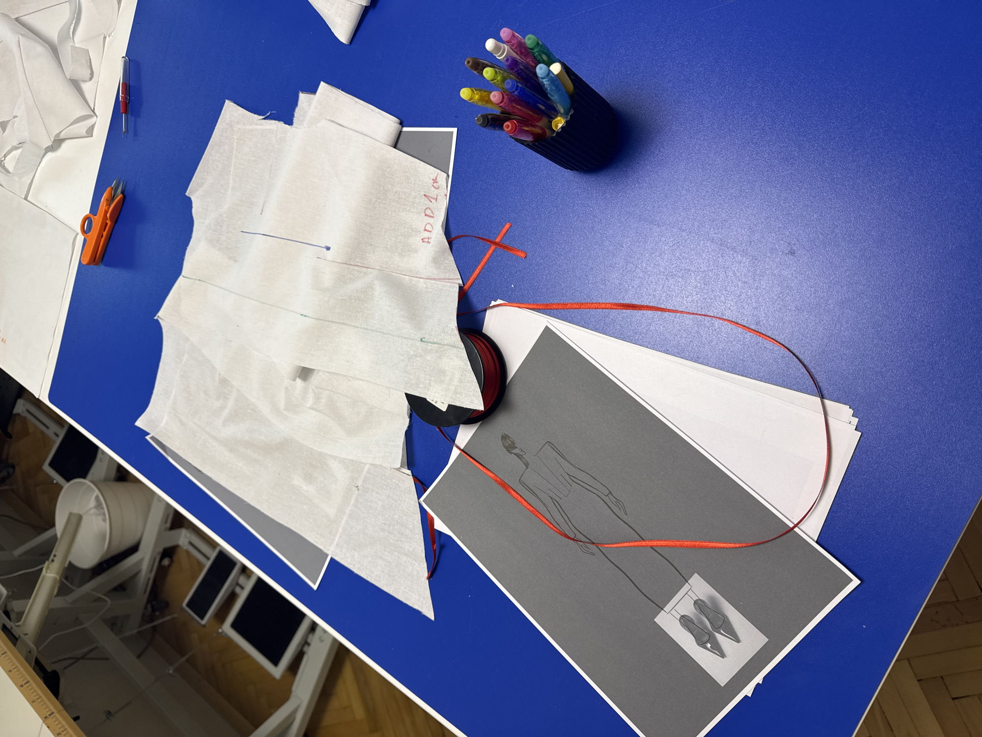 The scene depicts a fashion design or tailoring workspace located in Sofia, Bulgaria. The primary surface is a royal blue textured tabletop. In the center, several pieces of white translucent material, consistent with fabric patterns or muslin, are arranged. One piece visibly displays handwritten text "ADD 1cm" in red, along with colored marker lines. Adjacent to these materials, positioned towards the top right, are an orange plastic thread snip and a red-handled seam ripper.
In the lower left quadrant of the table, a stack of papers includes a top sheet of gray cardstock featuring a monochrome illustration of a tall, slender female figure in a long-sleeved dress, sketched in outline. Below the figure, a white rectangular inset on the gray card displays a detailed illustration of two pointed high-heeled shoes. A black spool of red ribbon rests on the table next to this stack, with a length of the ribbon extending across the illustration and onto the blue surface. To the right of the patterns and ribbon, a black corrugated cup holds an assortment of approximately a dozen colored markers and pens.
The background, partially visible on the left side of the image, shows a light wooden floor and the white metallic framework of what appears to be an industrial sewing machine table or cutting station, equipped with multiple black foot pedals and a white cylindrical waste bin. The overall arrangement suggests an ongoing process of garment design and creation.