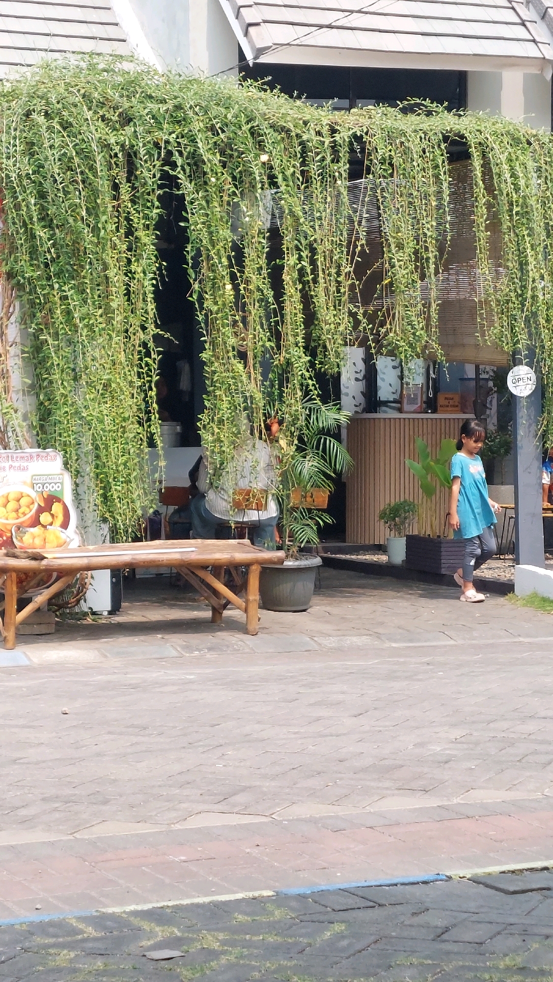 A young girl in a blue tunic and leggings walks past a cafe in Jimbaran Kulon, Indonesia. The cafe has an exterior draped with hanging plants. In the foreground, a rustic wooden bench sits on a paved walkway. Behind the bench is a sign advertising food. A person is seated inside the cafe. A potted palm tree stands to the right of the entrance. The sky is visible in the background.