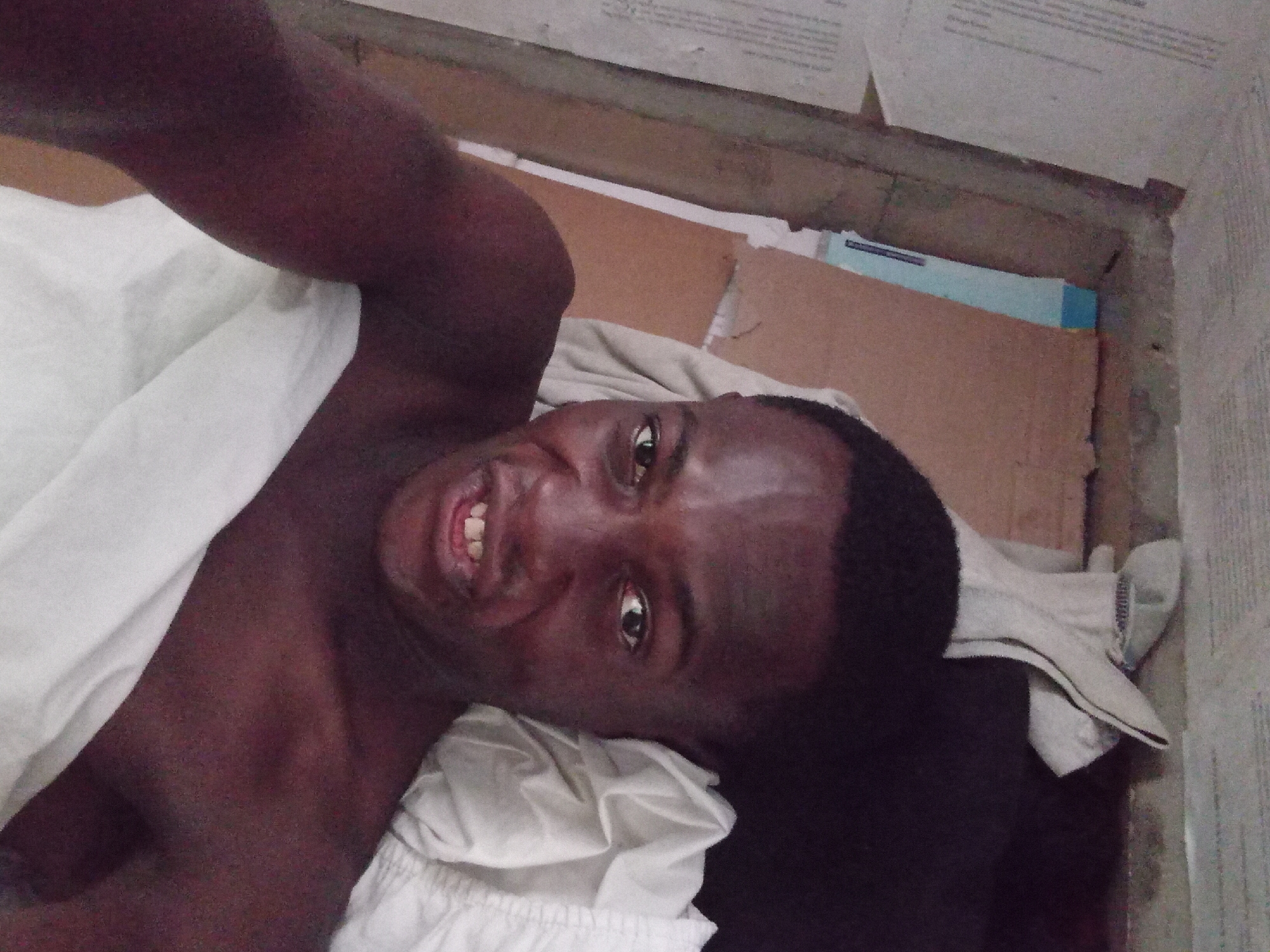 A man is lying down on a bed with white sheets, facing the camera. His head is tilted to the right. There is cardboard on the floor near the bed. The scene appears to be indoors. There is no information to indicate a location in Akwete, Nigeria.
