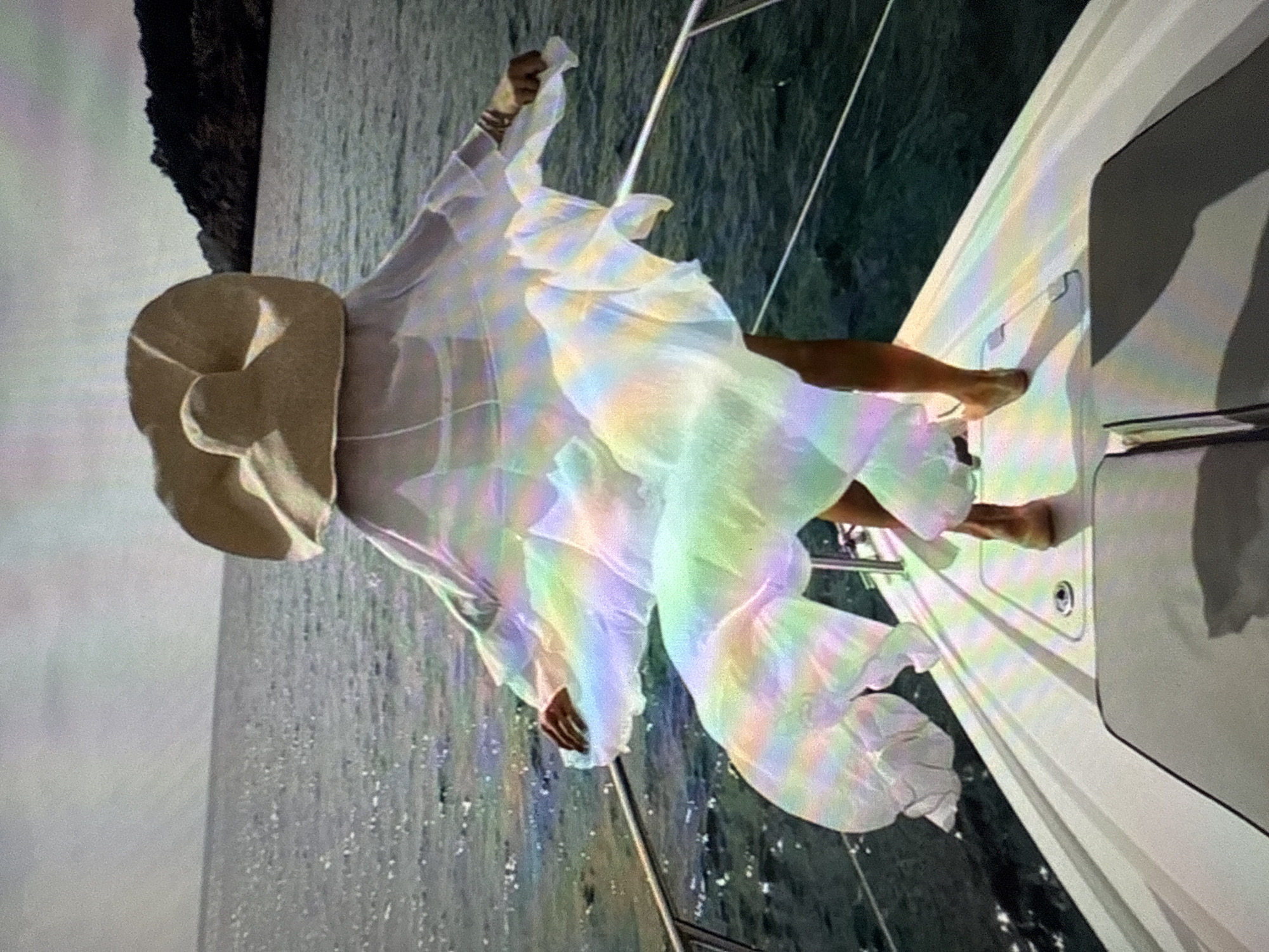 A person stands on the deck of a white boat, facing away from the camera, wearing a large, wide-brimmed straw hat and a sheer, flowing white garment that appears to be a cover-up or dress. The garment billows out behind them, catching the light and creating a rainbow effect. The person is wearing a bikini underneath the sheer garment. Their legs are visible, and they are barefoot. The boat is on the water, with the sea stretching out to the horizon under a bright, overcast sky. In the background, a dark, hilly coastline is visible. The image is a photograph, likely taken during the daytime. The location is not Găvănoasa, Moldova, but appears to be a coastal area.