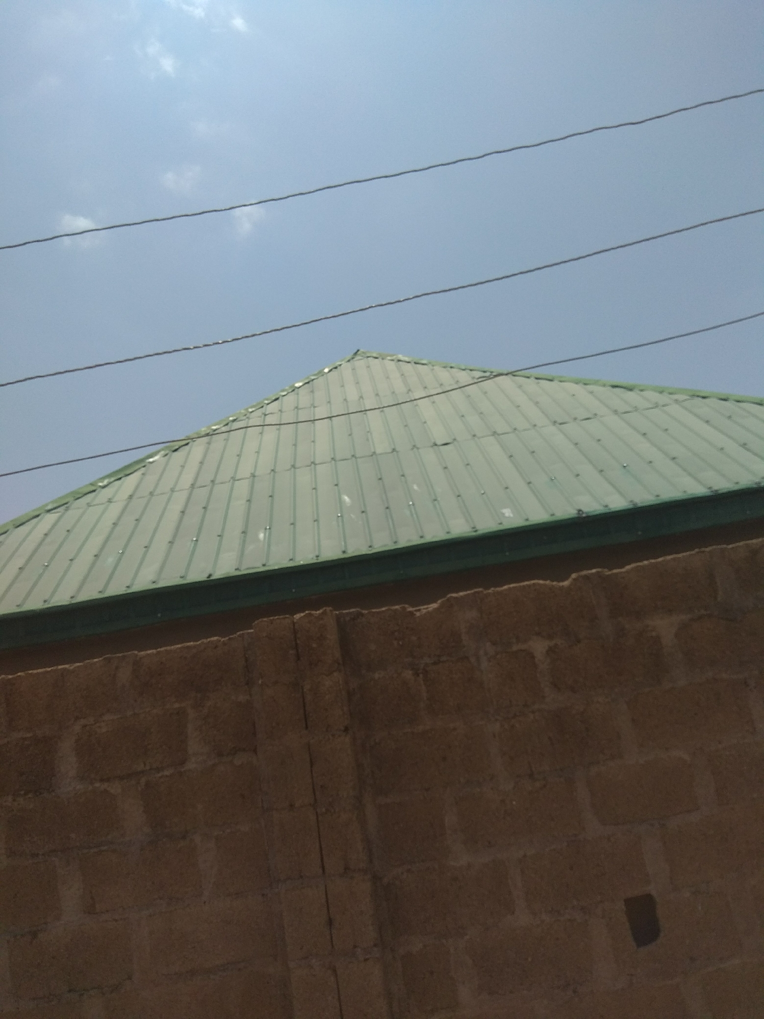The image shows a close-up view of a building's exterior, including a portion of its green, corrugated metal roof and a brown brick wall. The roof has a distinctive peak and is supported by visible beams underneath. Several power lines are strung across the sky, which is a pale blue with a few scattered clouds. The brick wall is constructed from rough, rectangular blocks. The scene appears to be an outdoor setting, likely a residential or commercial property, under a daytime sky. The specific location in Jalingo, Nigeria, is not visually identifiable from the image alone.