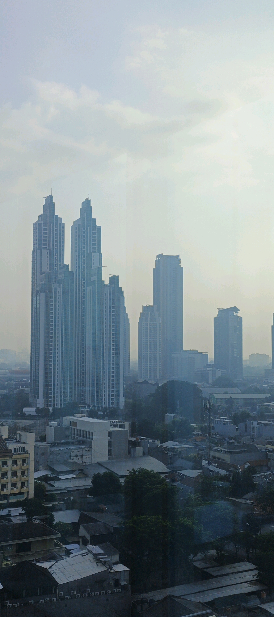 The image is an aerial view of a densely built urban environment in South Jakarta, Indonesia, during daylight hours. The scene is dominated by a cluster of modern high-rise buildings, characterized by their sleek architectural designs and glass facades, reflecting the muted sky. In the foreground and midground, lower-rise residential and commercial structures are visible, interspersed with pockets of trees and vegetation. The overall atmosphere appears hazy or overcast, with a pale sky suggesting either an early morning, late afternoon, or a day with atmospheric pollution. No individuals or specific actions are discernible in the image. The perspective suggests the photograph was taken from an elevated position, likely another building.