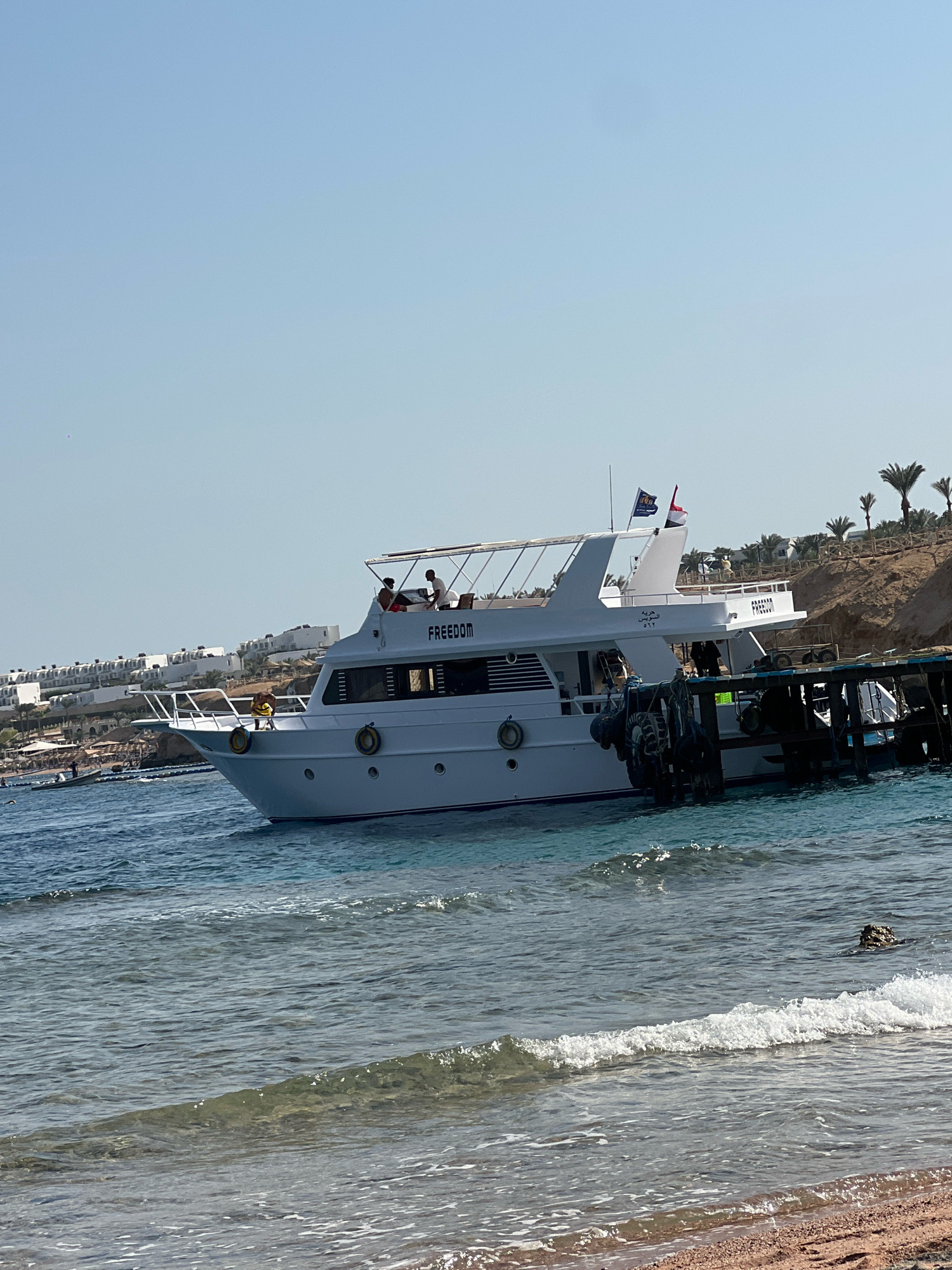 A white motor yacht named "FREEDOM" is docked next to a wooden pier. The yacht has several people on board, some visible on the deck. In the background, there is a beach with sun loungers and umbrellas, and buildings constructed on a hillside, characteristic of Sharm El Sheikh, Egypt. Palm trees are scattered along the coastline. The water is clear and blue, with small waves lapping at the shore in the foreground. The sky is clear and bright blue, suggesting a sunny day.