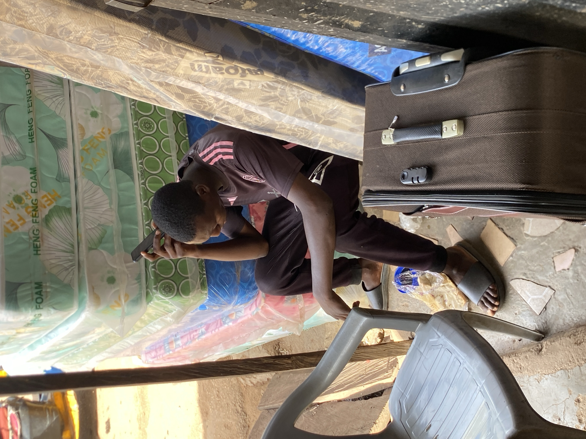 A male person is positioned in front of a stack of mattresses wrapped in transparent plastic. He is crouching and holding a black smartphone to his ear. He is wearing a dark-colored shirt with pink stripes, black pants, and dark-colored sandals. Beside him is a brown suitcase with a combination lock. The setting appears to be indoors, with the presence of a gray plastic chair in the foreground, and fragments of cardboard are visible on the ground. The context of "Bukuru, Nigeria" suggests this is a local business or storage area, but the image itself does not confirm this.