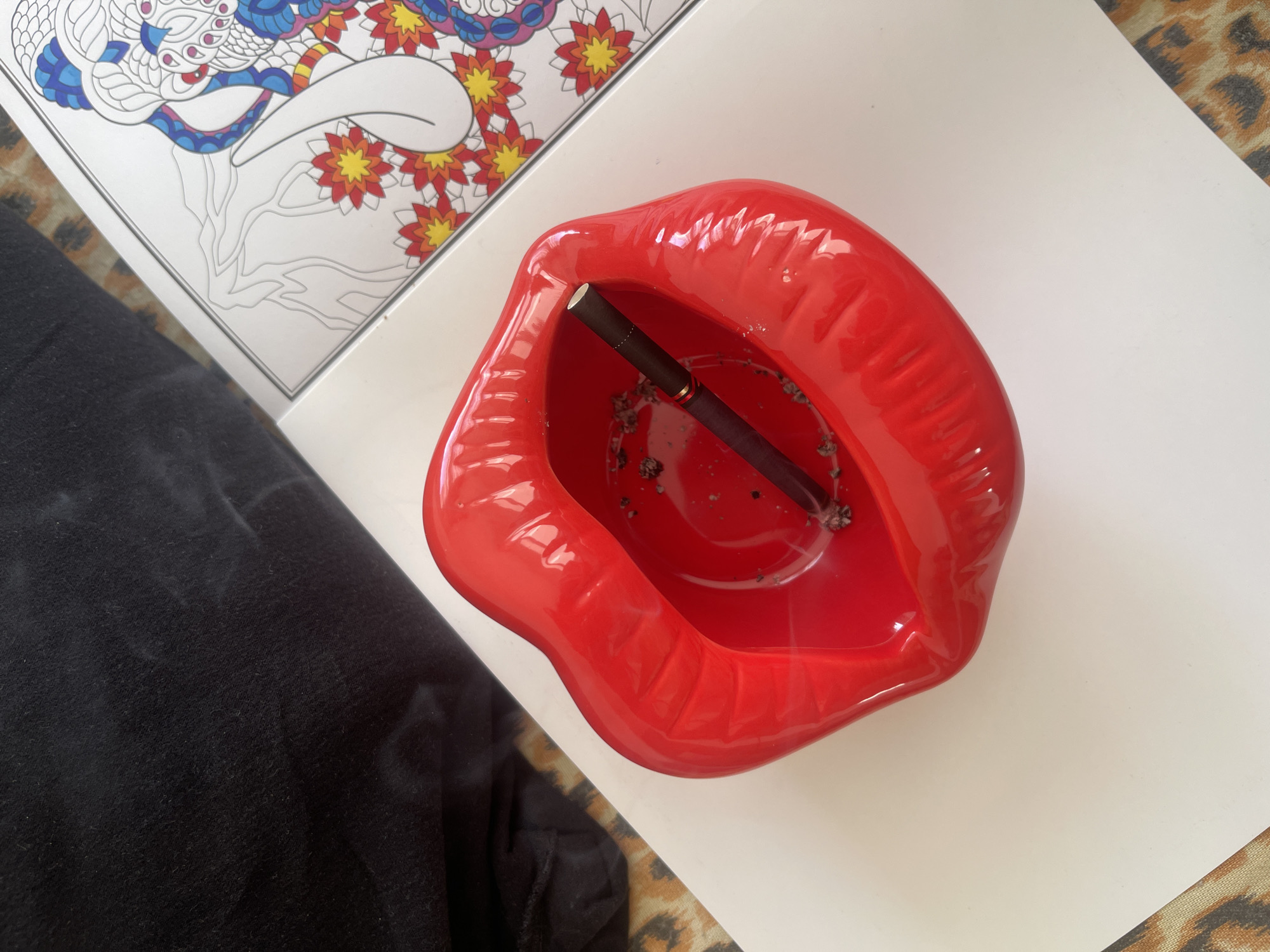 This is a high-angle, close-up shot of a red, lip-shaped ashtray containing a single cigarette with ash and a bit of smoke. The ashtray sits on a white surface, which appears to be a book with a coloring page of a detailed, colorful elephant. To the left of the ashtray is a black, textured fabric, likely clothing. The background shows a leopard-print pattern. The scene is indoors and does not provide any specific clues to its location in Varna, Bulgaria.