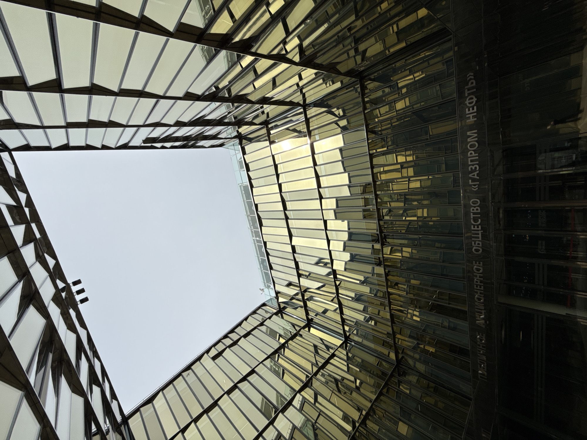 A low-angle shot shows the architecture of a building, looking up into the sky. The building's facade is composed of gold-tinted, rectangular glass panels framed by dark metal. The building's name is visible on its right side, written in Cyrillic: "ПУБЛИЧНОЕ АКЦИОНЕРНОЕ ОБЩЕСТВО «ГАЗПРОМ НЕФТЬ»". The sky is overcast and light gray, suggesting it may be cloudy. Three small lights are visible near the top left corner, on the building's facade. The architecture appears modern. This is the Gazprom Neft building in Saint Petersburg, Russia.