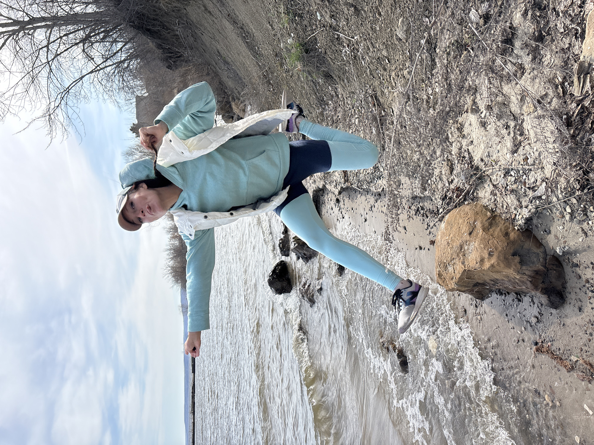 A young person, likely female, stands on the sandy and rocky bank of a large body of water, possibly a river or lake, in Ulyanovsk, Russia. She is dressed in activewear, featuring a light blue hooded sweatshirt, a white quilted vest, dark blue shorts worn over light blue leggings, a light-colored baseball cap, and athletic shoes with dark laces. She strikes a dynamic pose, with her left leg bent and foot lifted, and her arms raised with hands near her shoulders. She looks directly at the camera with a neutral expression.
Small waves are visible along the water's edge. The shoreline features pebbles, sand, and a prominent reddish-brown boulder to the person's right. In the background, under an overcast sky, are bare trees and distant land, suggesting a late winter or early spring setting. The scene is illuminated by soft, diffused daylight. No text is visible in the image.