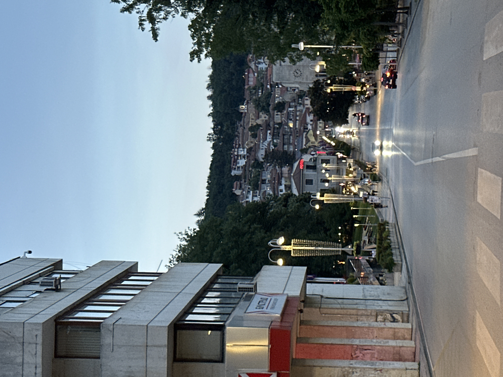 An evening urban landscape in Veliko Tarnovo, Bulgaria, is depicted at dusk, with artificial lights beginning to dominate the scene. The foreground features a modern building with a distinctive facade of concrete fins and large windows. A sign on this building clearly displays "BUMAG TREYDING" in Cyrillic, with additional red branding below.
To the right, a wide street is busy with traffic, illuminated by the bright headlights of approaching cars and the red taillights of those moving away. A crosswalk is visible in the bottom right corner. Along the street, a pedestrian area or park is lit by numerous decorative streetlights and lined with trees and bushes. People can be seen strolling or gathered in this area, suggesting a lively yet peaceful atmosphere. Further down the street, the word "HOTEL" is visible in red letters on a building.
In the background, a steep, forested hill rises, densely covered with traditional houses featuring red-tiled roofs, characteristic of Bulgarian architecture. A prominent building with a clock tower stands out among these historic structures, hinting at a central, older part of the town. The sky above remains a pale, clear blue, transitioning from day to night. The overall impression is one of a vibrant town beginning its evening activities.