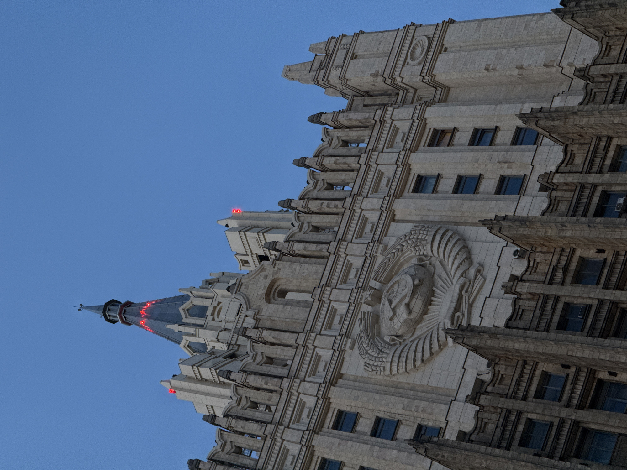 The image shows a portion of the Ministry of Foreign Affairs building, one of the Seven Sisters skyscrapers in Moscow, Russia. The architecture features a Stalinist Gothic style, with a prominent Soviet coat of arms on the facade. The building has a multi-tiered structure with numerous windows, decorative elements, and a spire at the top, which is illuminated with red lights. The sky is blue.
