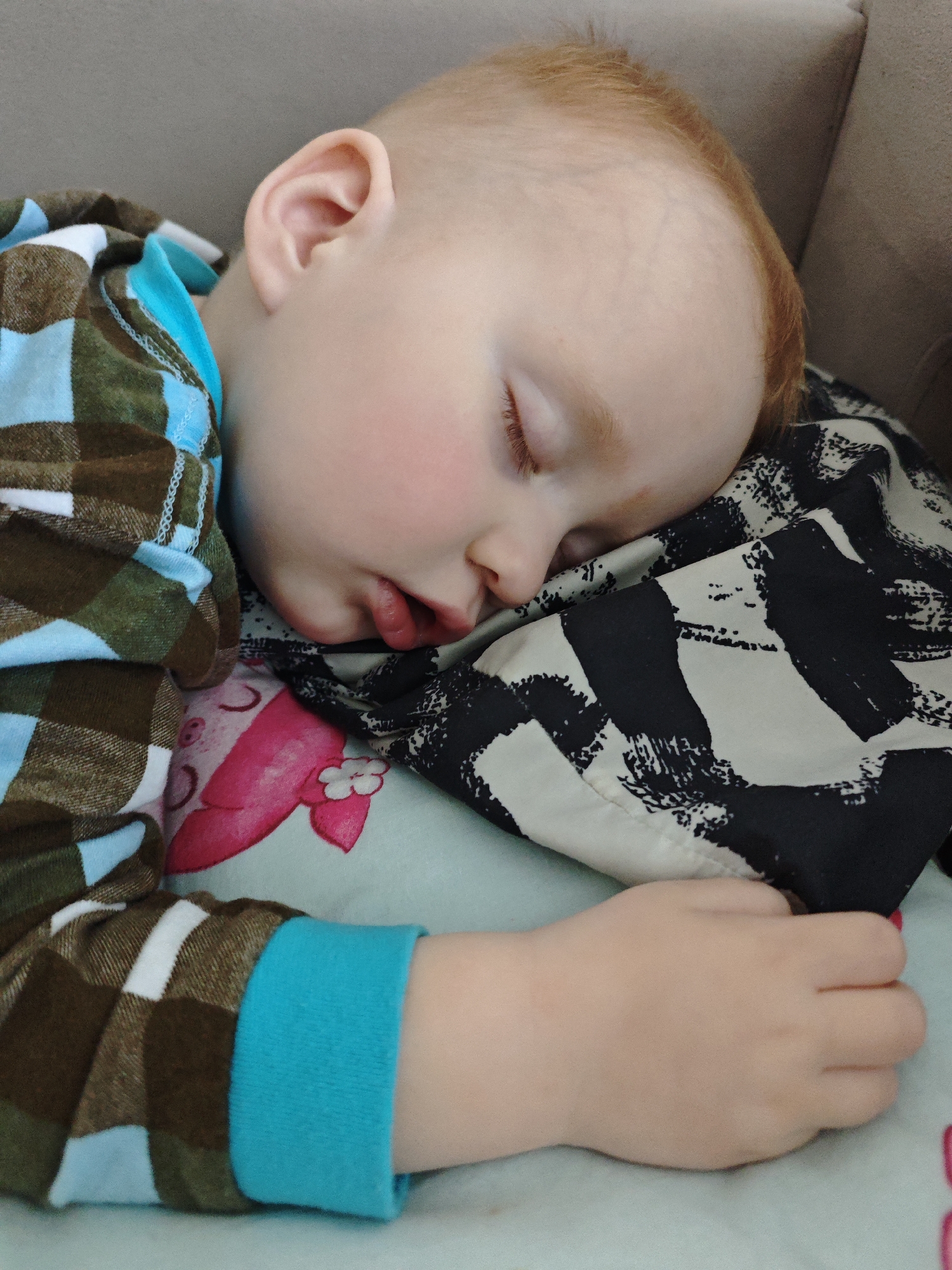 A fair-skinned, red-haired toddler sleeps peacefully on a pillow. The child is wearing a patterned pajama top with blue cuffs and a hood. The pillow beneath the child’s head has a black and white pattern on top of a pale blue base. Part of a cartoonish pink flower is also visible on the pale blue pillow. The toddler’s hand is curled in a fist, resting near the pillow. The setting appears to be indoors on a sofa or bed. There is no indication of this photo's location being in Markovskii, Russia.