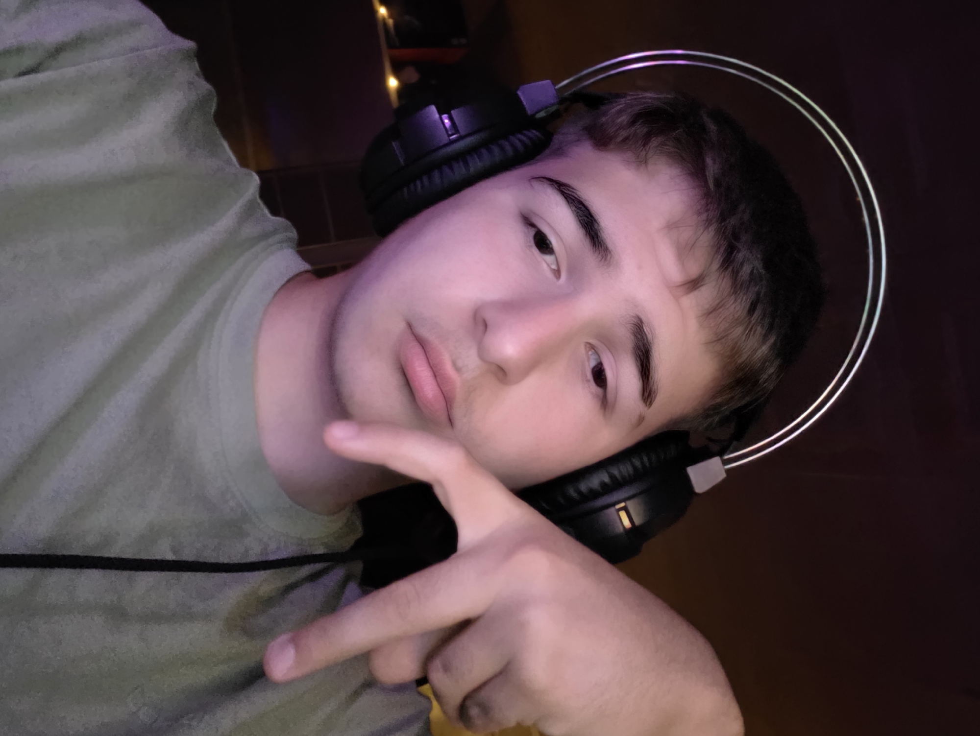 A young person with dark hair and headphones is looking down and to their left. Their right hand is raised, with the index and middle fingers extended in a "V" shape. They are wearing a green t-shirt. The background is dark with some purple and pink lighting, possibly from LED strips. The image appears to be a selfie taken indoors. There is no indication of the location in Aleksandrovo, Bulgaria.