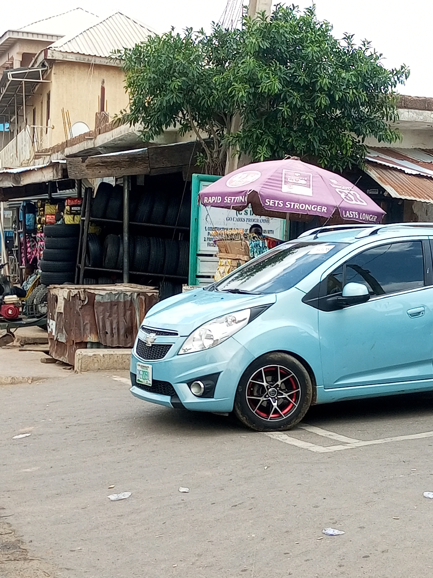 In Arawa, Nigeria, a light blue Chevrolet Spark is parked on a paved road in front of a roadside stall. The stall features stacks of tires, some bags, and an open umbrella advertising "RAPID SETTING SETS STRONGER LASTS LONGER". A person is partially visible behind the counter, wearing a patterned top and head covering. The building behind the stall has multiple levels with a corrugated metal roof. The car has black alloy wheels with red accents. The ground is littered with small pieces of paper and debris. The scene appears to be a commercial area with various goods for sale.