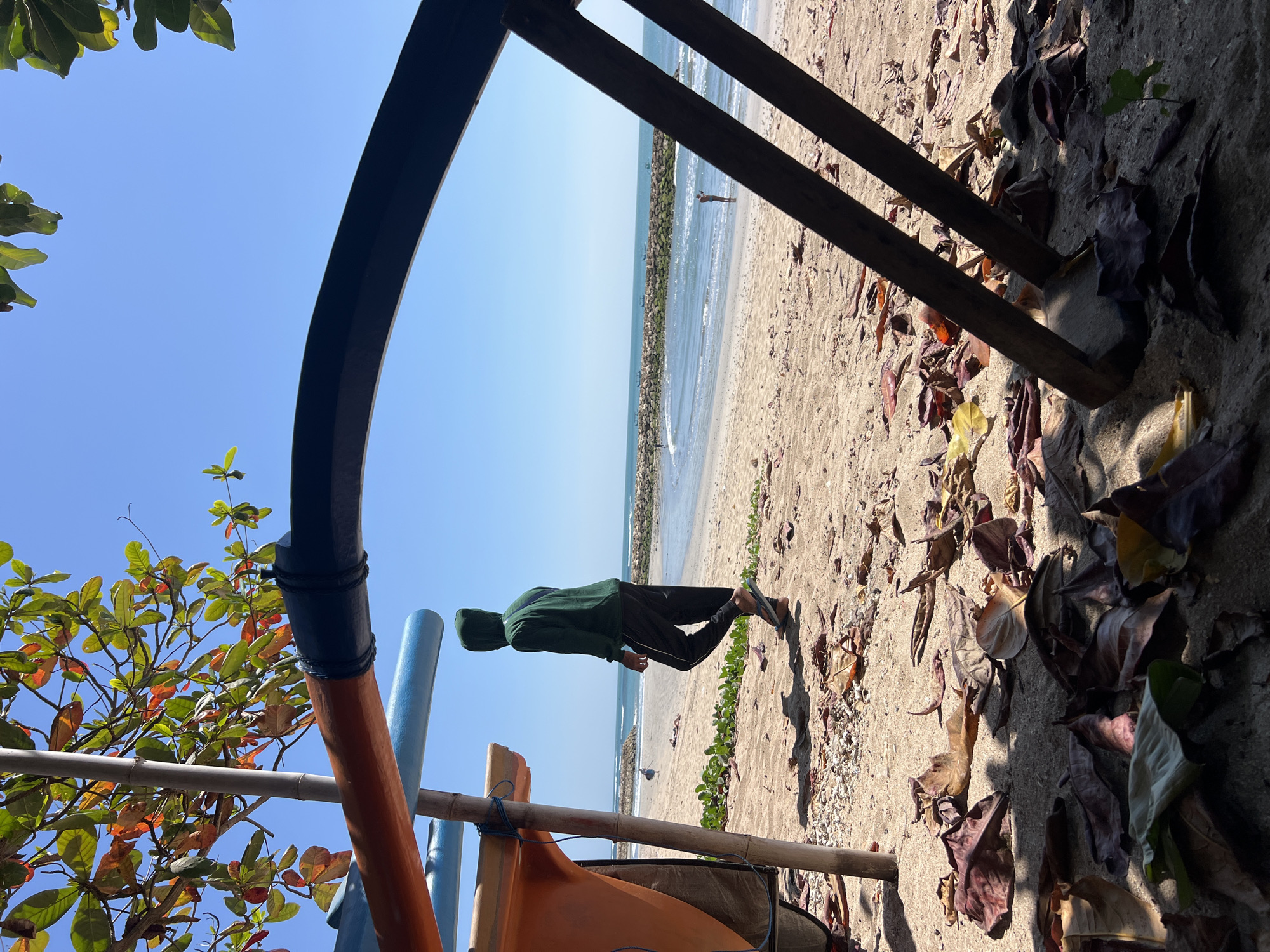A person wearing a green hoodie and black pants walks on a sandy beach in Kuta, Indonesia, with the ocean in the background. In the foreground, there are fallen leaves scattered on the sand, and part of a boat is visible. The sky is clear and blue, and sunlight casts shadows on the beach.