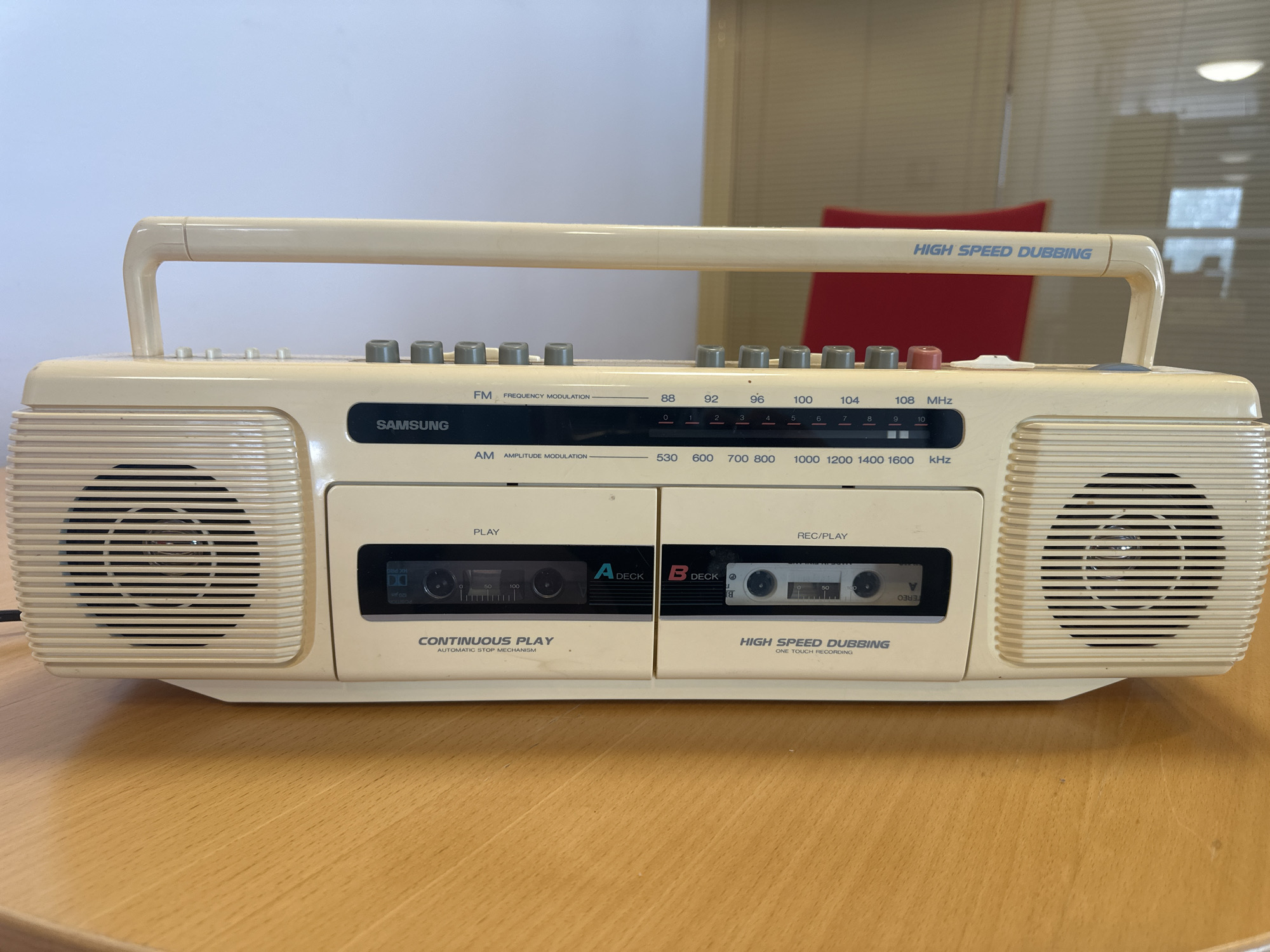 The image shows a close-up, eye-level shot of a beige Samsung boombox with a handle, placed on a wooden table. The boombox features two cassette decks, one labeled "A DECK" and the other "B DECK", with visible cassette tapes inside. Above the cassette decks, there is a radio tuner with FM and AM frequency displays and tuning knobs. The words "HIGH SPEED DUBBING" are printed on the top right of the boombox. The background is out of focus, showing a light-colored wall and vertical blinds, with a hint of a red chair. The scene appears to be indoors. No people are visible in the image, and no specific location in Espoo, Finland can be identified.