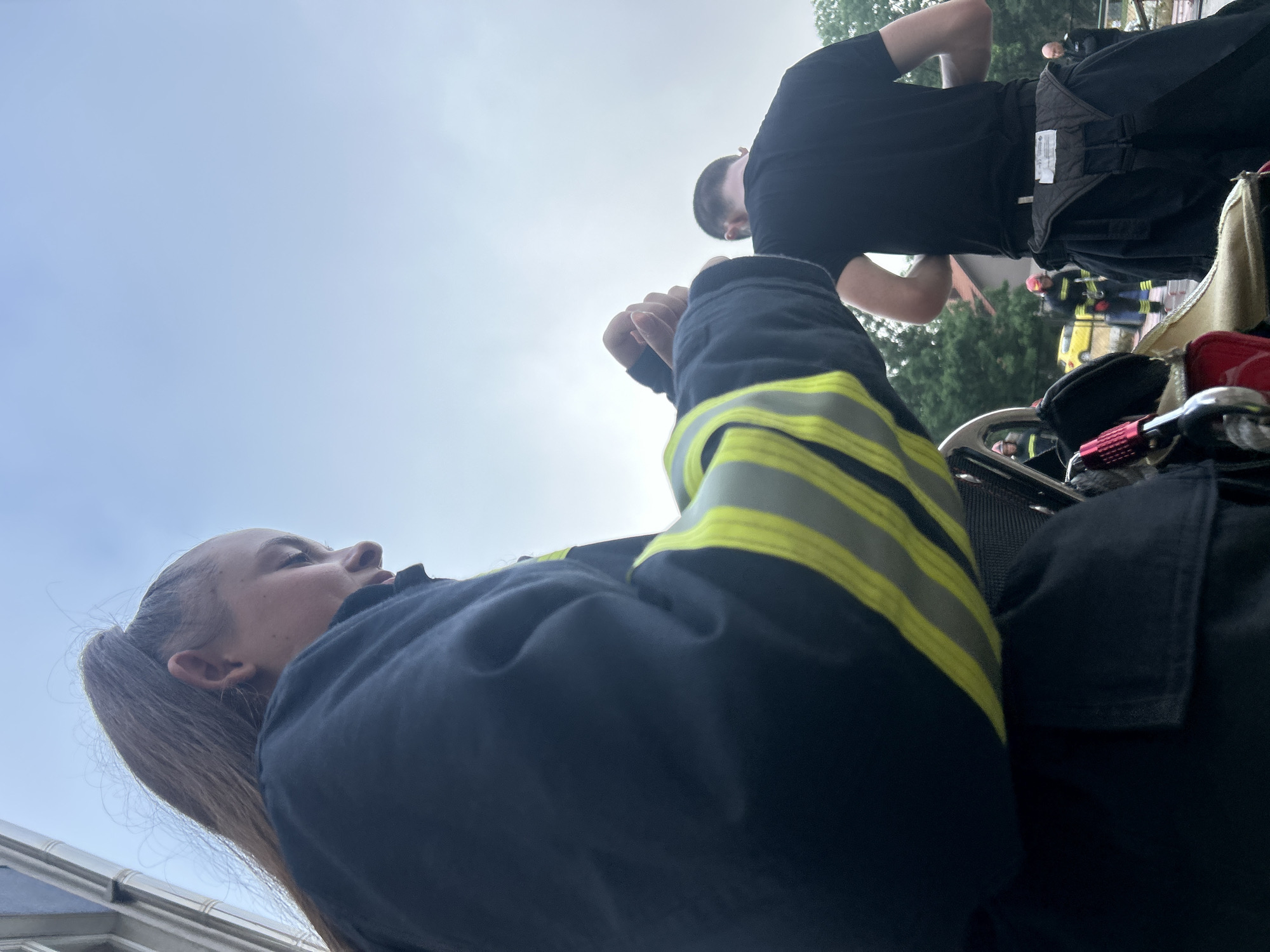 The media file presents a low-angle shot of an outdoor scene, rotated so subjects appear to be leaning or lying down, but conceptually captured looking upwards at people and the sky.
In the foreground, a young female is visible, seen from the chest up on the left side of the frame. She has light brown hair pulled back in a ponytail and is wearing a dark blue uniform jacket with prominent reflective yellow stripes on the arm, characteristic of emergency services such as firefighters. Her hands are clasped, and she is looking upwards and slightly to the right with a neutral, observant expression.
In the mid-ground, another individual is seen from behind, wearing a black t-shirt and dark pants with a utility belt. Further in the background, several other figures are discernible, some appearing to be in similar emergency uniforms, including one with a red helmet. A yellow vehicle is partially visible among the background elements.
The setting is outdoors, with a bright, likely overcast sky filling the upper portion of the frame, indicating daytime. Trees and glimpses of buildings suggest an urban or semi-urban environment. Various pieces of equipment, including red and metallic components, are visible near the foreground subject, reinforcing the context of an emergency services operation or training event. There is unidentifiable text on a white label attached to the utility belt of the person in the black t-shirt. The overall atmosphere is one of focused activity or observation by emergency personnel, consistent with the location context of Sofia, Bulgaria.