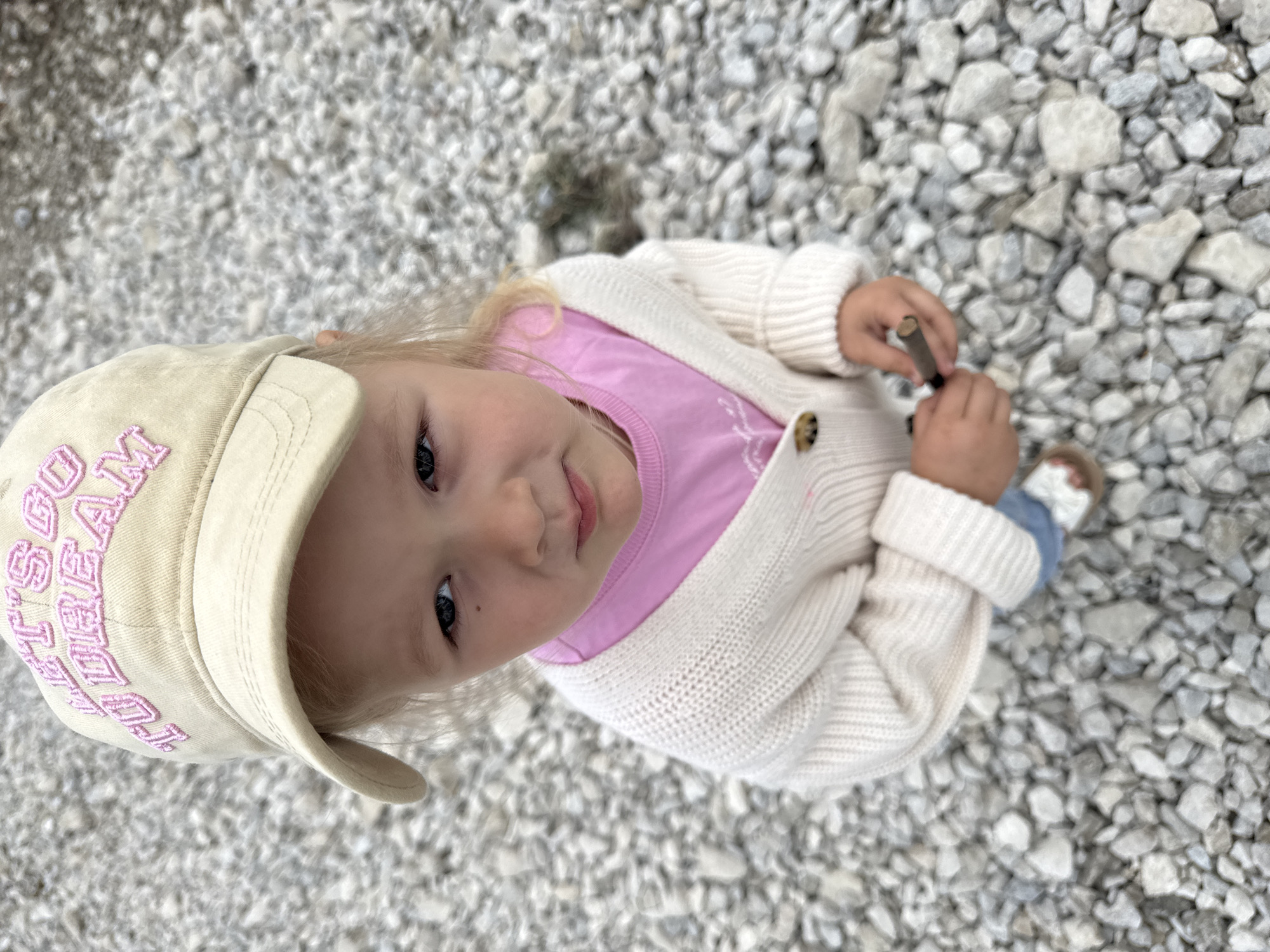 This media file features a close-up, high-angle shot of a young child lying on a bed of gravel. The child, with blonde hair and fair skin, is wearing a pink t-shirt, a cream-colored knitted cardigan, and a beige baseball cap. The cap has pink embroidered text on it. The child is holding a small, dark-colored object in their hands, possibly a twig or a small toy. The child's expression is a gentle smile, and their eyes are looking upwards towards the camera. The background consists entirely of gravel, with stones of various sizes and colors, predominantly white and gray. The lighting suggests it could be daytime, possibly outdoors given the gravel setting. No specific weather cues are evident, but the overall mood appears calm and serene. The location context provided is Горный, Russia.