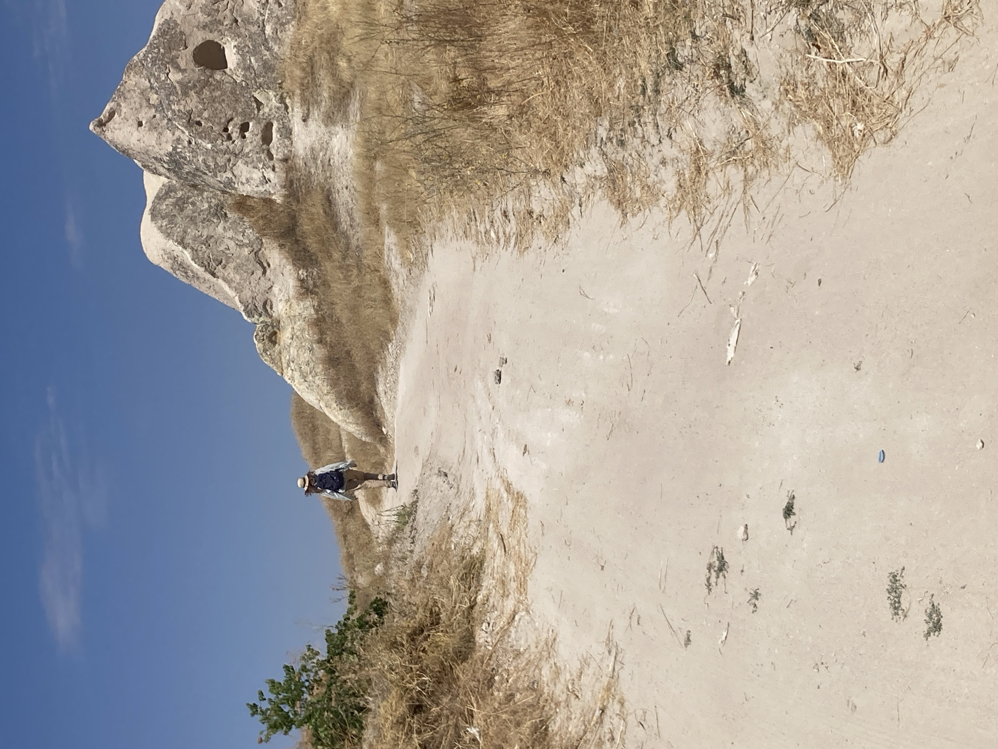A lone hiker wearing a straw hat and a backpack walks up a dusty, sandy path in Göreme, Turkey. The landscape is arid, with dry, golden grass lining the path and unusual rock formations in the background. One of these formations has natural holes, suggesting it might be a cave dwelling or natural erosion. The sky is a clear, bright blue.