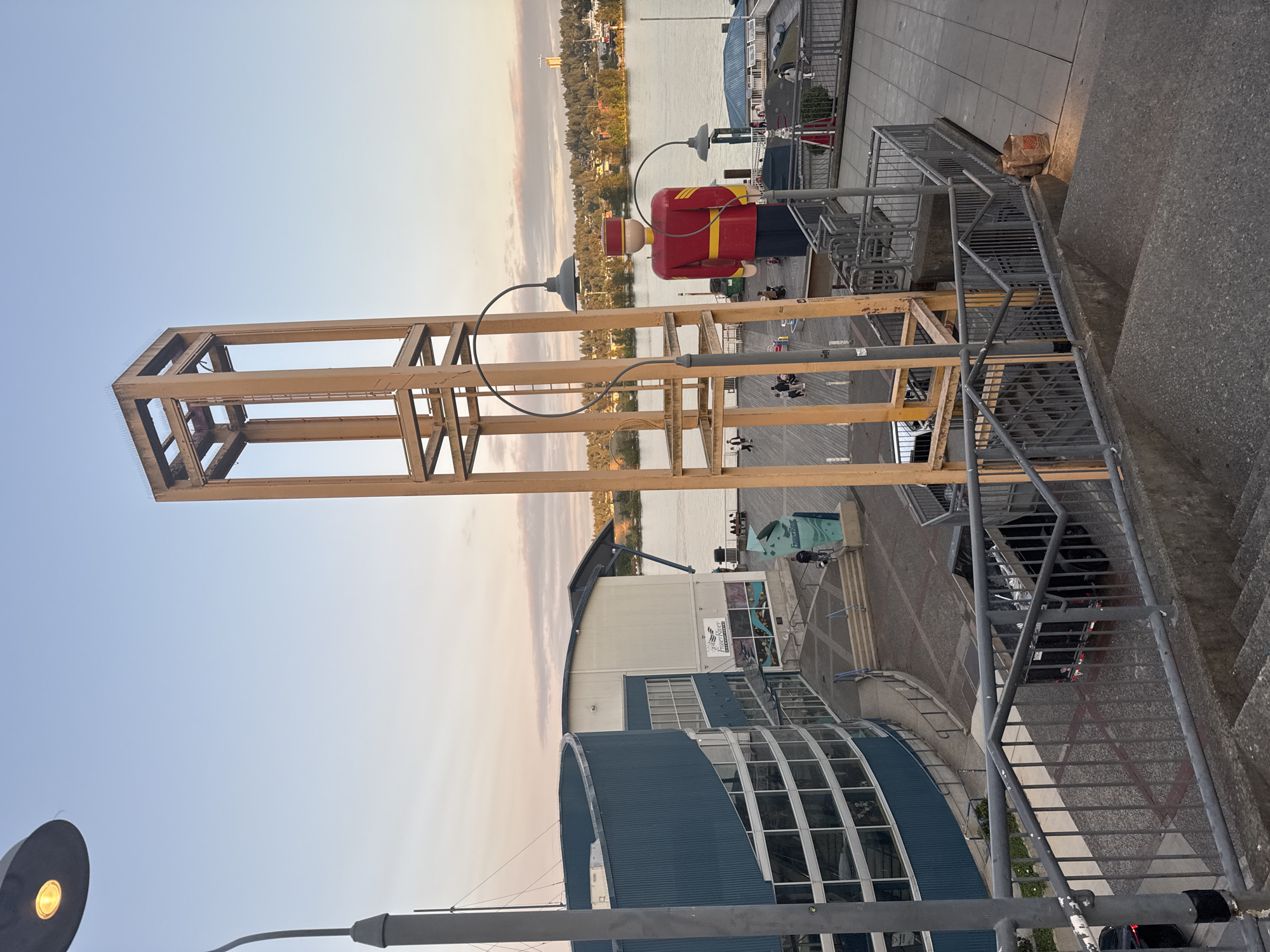 The image depicts the exterior of the Fraser River Discovery Centre in New Westminster, Canada, at dusk. A large, brightly colored statue of a soldier in a red uniform stands prominently on a wooden pier overlooking the Fraser River. In the foreground, a concrete staircase leads up to the viewing area. A modern, curved building with blue siding and large windows, identified as the Discovery Centre, is visible to the left. A tall, open-air metal structure, likely a clock tower or observation point, is also present. Several people are seen walking on the pier and near the entrance of the building. The sky is a soft gradient of blue and pink, indicating the setting sun. A black truck with a trailer is parked near the staircase in the lower right portion of the image.